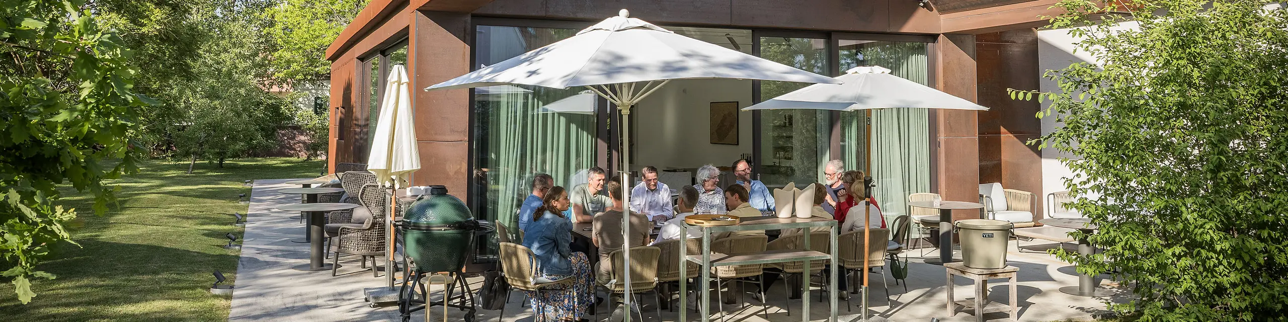 PREFARENZEN 2027 Dialóg odborníkov na terase modernej vinárskej rezidencie: Členovia poroty sedia okolo veľkého stola, intenzívne diskutujú o architektonických projektoch a listujú v knihách o architektúre. Scénu dotvára prirodzené svetlo a zelená záhrada, pričom na streche a fasáde budovy sú viditeľné hliníkové výrobky PREFA. Obrázok vyjadruje kreatívnu diskusiu, sústredenú analýzu a živú výmenu názorov. Strechy a fasády PREFA predstavujú inovatívne, trvácne a esteticky náročné riešenia, ktoré spájajú dizajn, funkčnosť a inšpiráciu v architektonických projektoch.