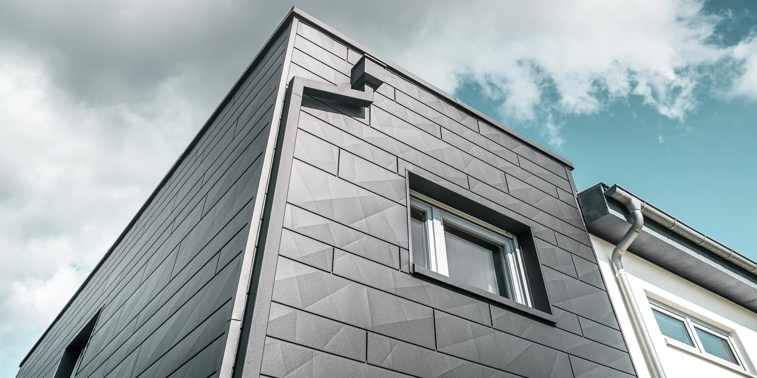 Výrazná fasáda zrekonštruovaného rodinného domu v Teuschnitzi je pokrytá hliníkovými panelmi PREFA Siding.X v čiernej farbe P.10, ktoré vytvárajú modernú a geometrickú štruktúru. Fasádne prvky majú jasné línie a zdôrazňujú nadčasový charakter budovy. Okno je orámované čiernymi hliníkovými kompozitnými doskami. Integrovaný odvodňovací systém PREFA s hranatými zvodmi a zbernou nádržou na vodu sa harmonicky začleňuje do budovy a poskytuje štýlové a funkčné riešenie.  Translated with DeepL.com (free version)