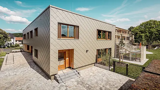 Obrázok zobrazuje materskú školu v Alzenau zo šikmej vtáčej perspektívy a zdôrazňuje modernú architektúru s výraznou bronzovou hliníkovou fasádou PREFA. Fasáda je tvorená fasádnymi šablónami 44 × 44, ktoré vytvárajú harmonický šupinový vzor a dodávajú stavebnému objemu elegantnú dynamiku. Veľké drevené okná s vertikálnymi lamelami prinášajú teplé akcenty a prepúšťajú do interiéru množstvo denného svetla. Na pravej strane obrázka sa otvára areál s oploteným ihriskom, zelenými plochami a krytou terasou. Kombinácia trvácneho, bezúdržbového hliníka a udržateľných materiálov robí z budovy vzorový projekt funkčnej a ekologickej architektúry v oblasti vzdelávacích stavieb.