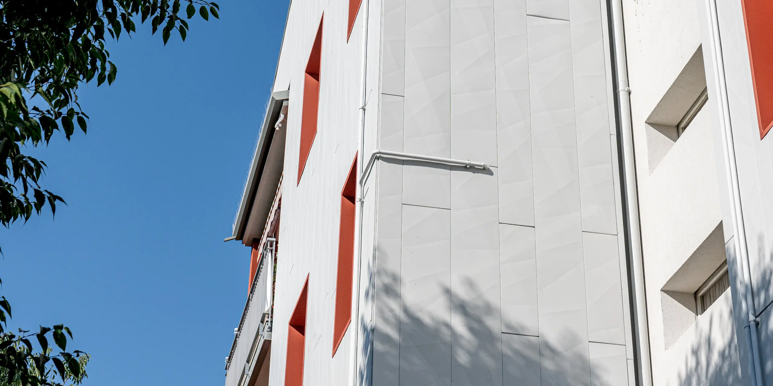 Die Aufnahme zeigt die modernisierte Wohnanlage Alba B aus einer dynamischen Untersicht, eingerahmt von grünen Baumästen und einem klaren blauen Himmel. Die helle, großflächige Fassade aus PREFA Siding.X Aluminiumfassadenpaneelen wirkt ruhig und zugleich lebendig: Feine Kantungen und die vertikale Verlegung erzeugen subtile Licht- und Schattenspiele. Auffällig sind die tief eingeschnittenen Fensteröffnungen mit kräftig roten Laibungen, die starke Kontraste setzen. Die langlebigen, witterungsbeständigen und wartungsarmen PREFA Aluminiumprodukte verbinden hier markante Architektur mit dauerhaftem Fassadenschutz.
