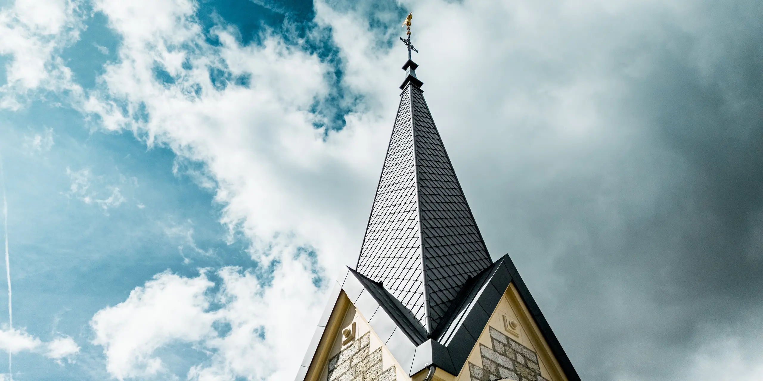 Spektakuläre Nahaufnahme der sanierten Kirchturmspitze in Le Sentier, Schweiz. Die PREFA Dachraute 29 × 29 in P.10 Dunkelgrau verleiht dem schlanken Turm eine elegante, präzise Struktur und setzt sich eindrucksvoll vor dem Wolkenhimmel ab. Die langlebige Aluminiumdeckung überzeugt durch hohe Witterungsbeständigkeit, geringes Gewicht und optimale Eignung für anspruchsvolle, steile Dachgeometrien.