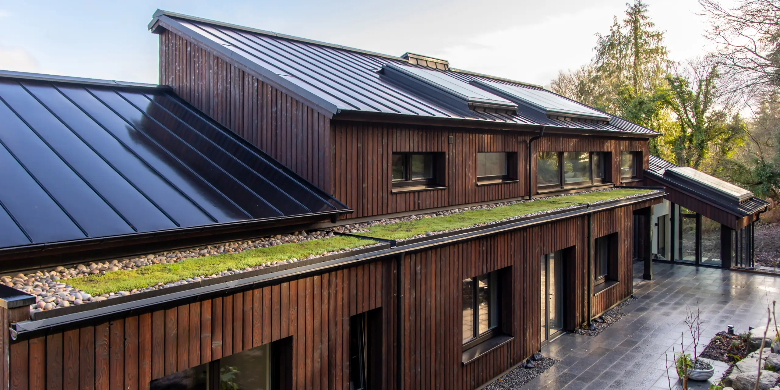 Schrägperspektive entlang der Terrasse des House of Holywell in Sligo: Die dynamisch verlaufenden Dachflächen aus PREFALZ Aluminium in P.10 Schwarz ziehen den Blick über die gesamte Gebäudelänge. Die präzisen Stehfalze strukturieren die Oberfläche und reflektieren das Licht, wodurch die klare Architektur zusätzlich betont wird. Die Kombination aus dunklem Dach, Holzfassade und begrünten Elementen schafft ein spannendes Gesamtbild. PREFA Aluminium überzeugt dabei durch Langlebigkeit, Korrosionsbeständigkeit und zuverlässigen Schutz bei jedem Wetter.