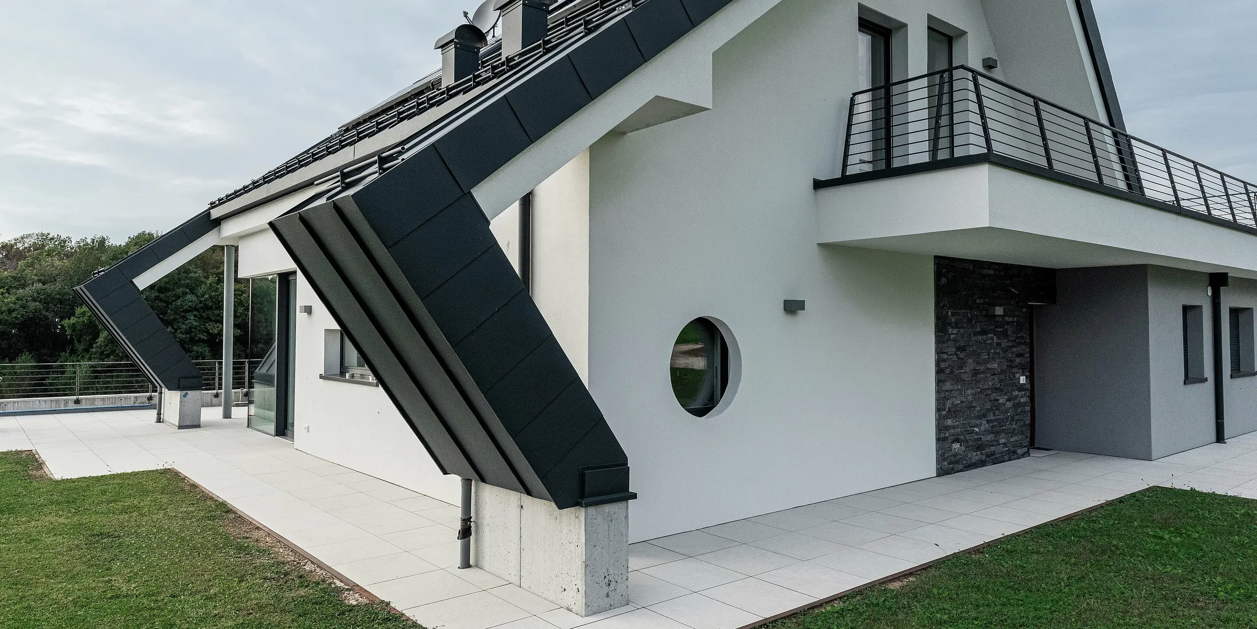 Detailný záber na šikmú strešnú nadstavbu moderného rodinného domu v Borgo Valbelluna s krytinou PREFALZ so stojatou drážkou vo farbe P.10 antracit. Nápadný presah hliníkovej strechy zdôrazňuje inovatívnu architektúru a chráni fasádu. Trvanlivá strešná krytina odolná voči poveternostným vplyvom dokonale ladí s bielou omietnutou fasádou a tmavými akcentmi. Obvodový balkón a okrúhle okenné detaily dodávajú budove osobitý nádych. Vysokokvalitný hliník PREFA zaručuje stabilitu, eleganciu a oslobodenie od údržby.