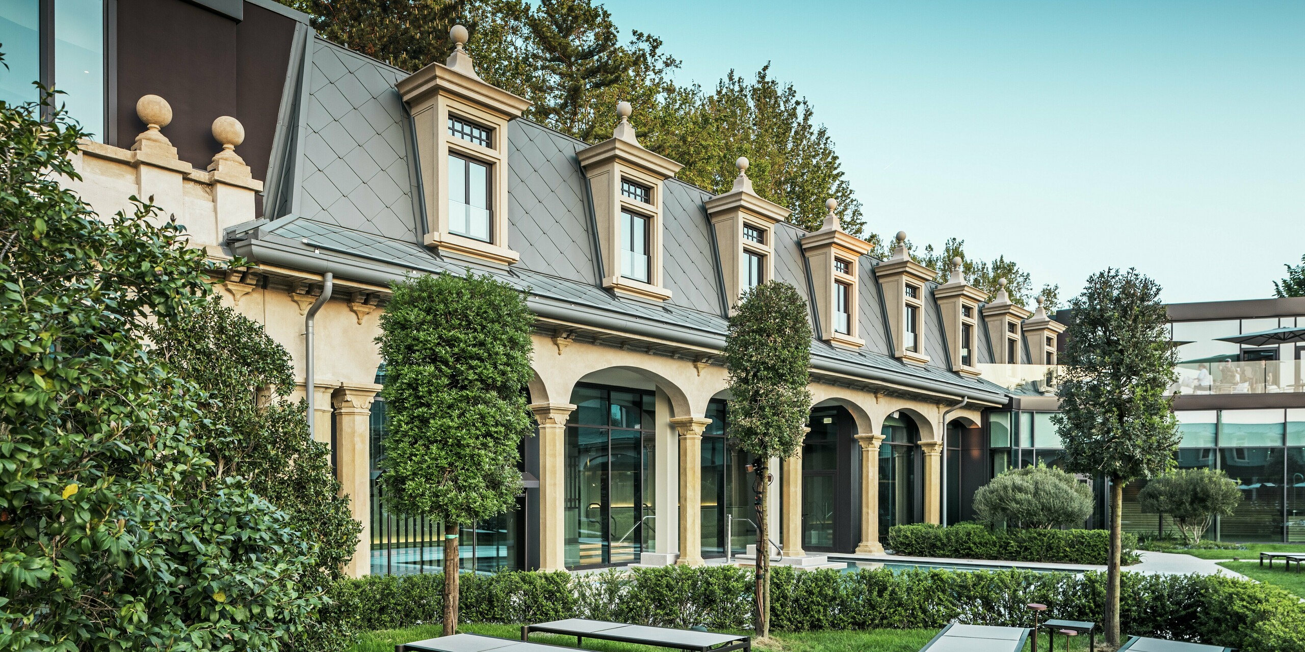 Detailný pohľad na štýlovo zrekonštruovanú strešnú krajinu termálneho kúpeľa „De Montel – Terme Milano“ s PREFA strešnými šablónami 44 × 44 v odtieni P.10 Svetlošedá. Presne vyhotovené vikiery, okenné rámy a rímsy podčiarkujú historickú eleganciu secesného komplexu. V popredí sa nachádza pokojná záhradná plocha s modernými ležadlami, geometricky strihanými živými plotmi a tvarovo krásnymi stromami – miesto oddychu s architektonickou hĺbkou.