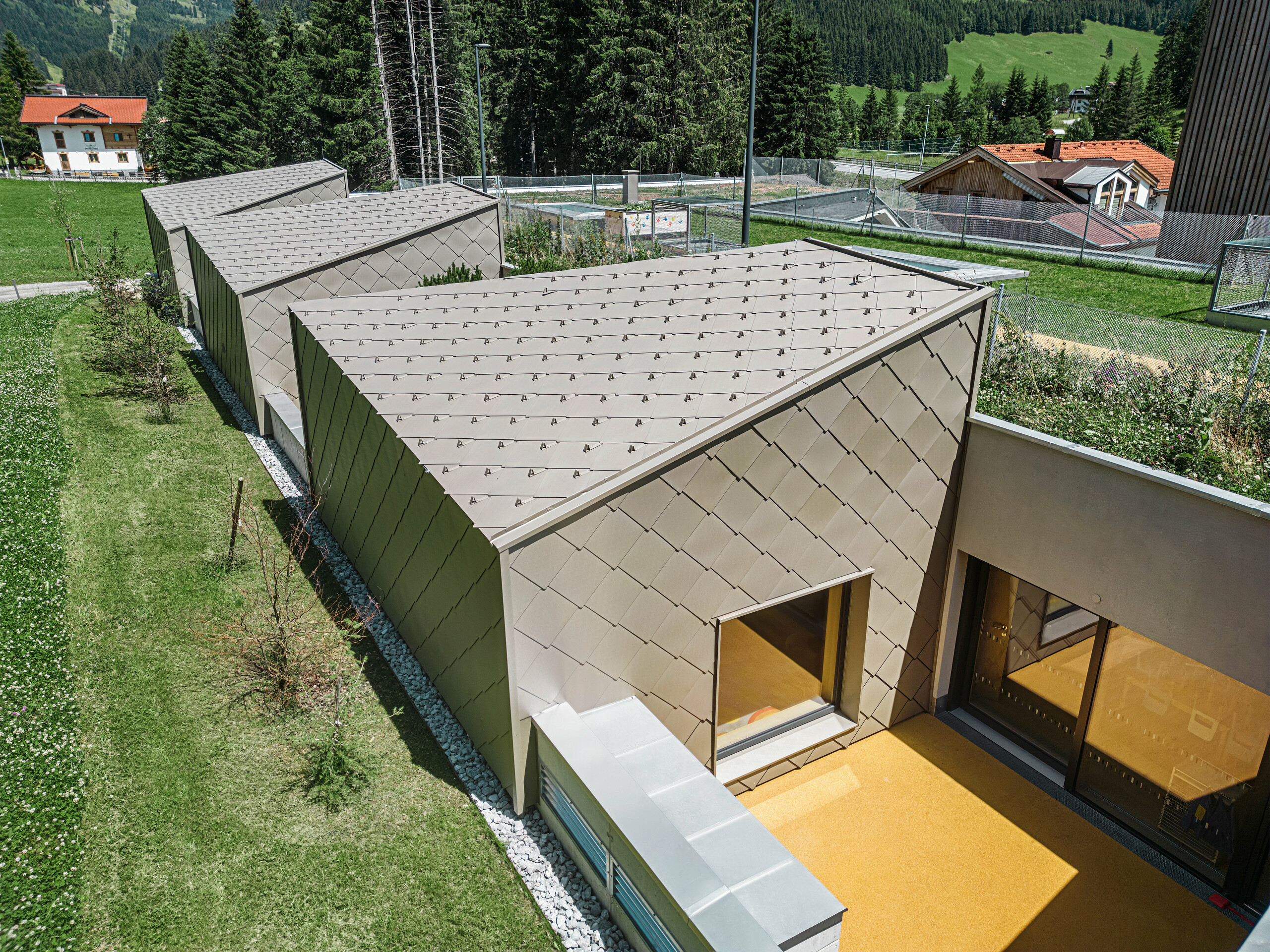 Bronzovo sfarbené strešné a fasádne šablóny PREFA 44 × 44 pokrývajú strechu a časť fasády modernej budovy v Gerlose v Zillertali. Budova má veľkorysé okenné priečelia a terasové plochy. V pozadí je vidieť vidiecke okolie so zelenými lúkami a zalesneným kopcom.