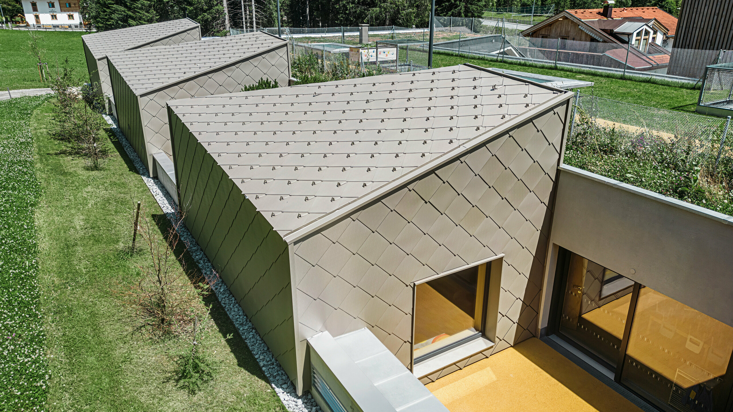 Bronzovo sfarbené strešné a fasádne šablóny PREFA 44 × 44 pokrývajú strechu a časť fasády modernej budovy v Gerlose v Zillertali. Budova má veľkorysé okenné priečelia a terasové plochy. V pozadí je vidieť vidiecke okolie so zelenými lúkami a zalesneným kopcom.