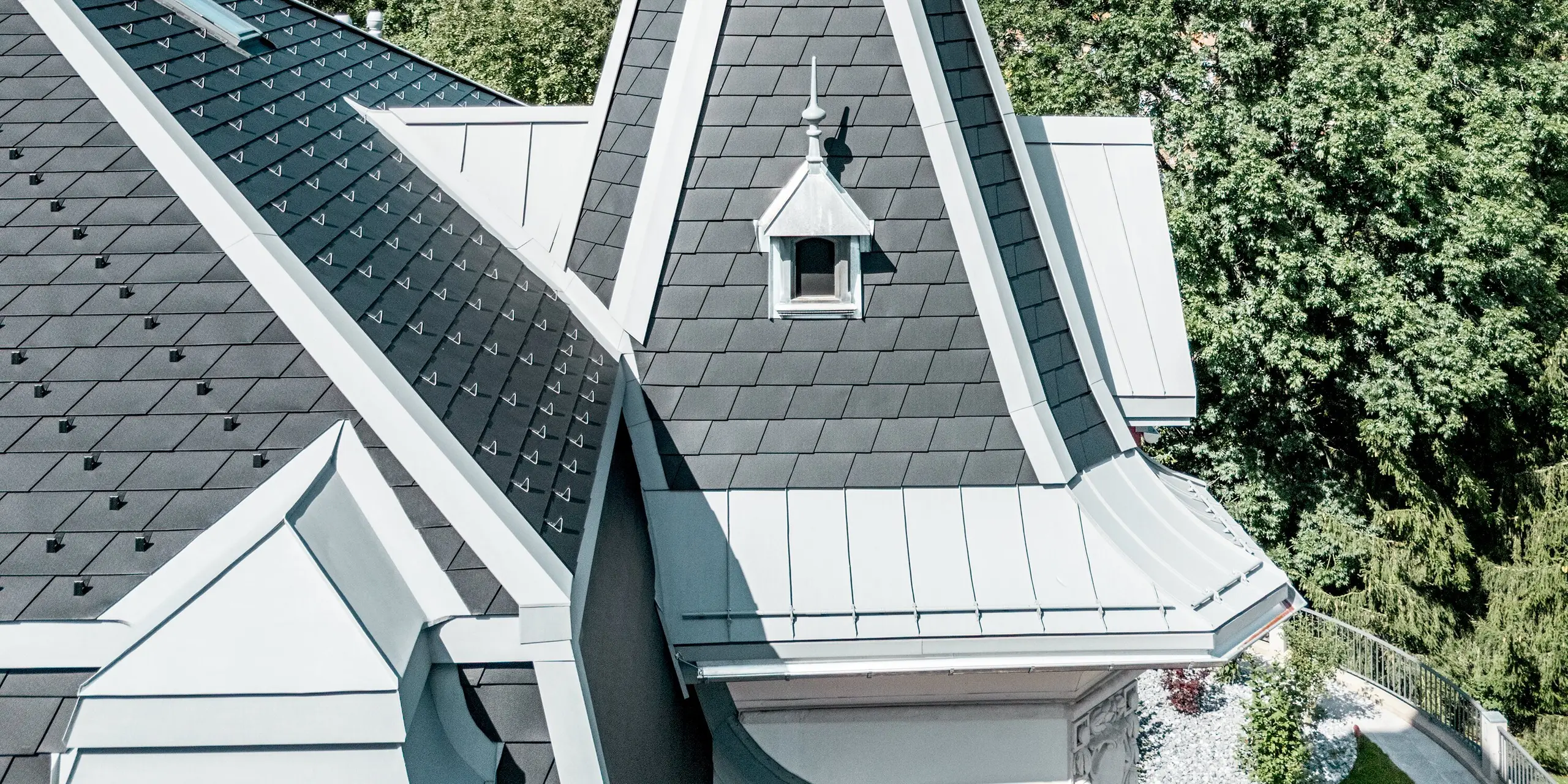 Detailaufnahme des filigranen Turms der Manufacture Horlogère du Locle in der Schweiz, der als eleganter Eyecatcher über der komplexen Dachlandschaft thront. Die steilen, präzise gedeckten Flächen aus PREFA Dachschindeln in P.10 Schwarz erzeugen eine markante Struktur, während PREFALZ in Patinagrau geschwungene Übergänge und feine Rundungen perfekt formt. Das leichte Aluminiumdach überzeugt durch hohe Witterungsbeständigkeit, außergewöhnliche Langlebigkeit und maximale Gestaltungsfreiheit für anspruchsvolle, architektonisch einzigartige Dachlösungen.