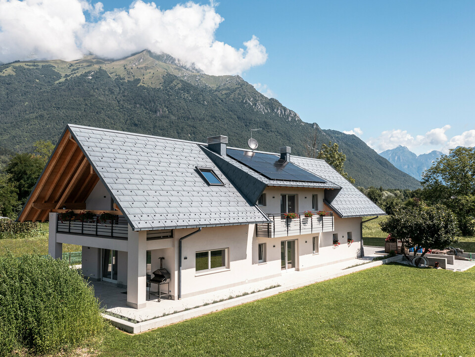 Záber z dronu na dom v talianskom Belluno, pokrytý strešnými panelmi PREFA R.16 vo farbe P.10 antracit. Nachádza sa v úchvatnej alpskej krajine a spája tradičné prvky, ako sú odhalené drevené štíty, s modernými, odolnými stavebnými materiálmi, čím vytvára dokonalú symbiózu tradície a modernosti.