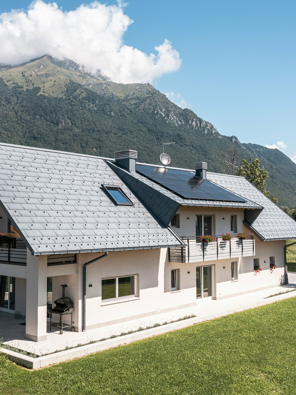 Záber z dronu na dom v talianskom Belluno, pokrytý strešnými panelmi PREFA R.16 vo farbe P.10 antracit. Nachádza sa v úchvatnej alpskej krajine a spája tradičné prvky, ako sú odhalené drevené štíty, s modernými, odolnými stavebnými materiálmi, čím vytvára dokonalú symbiózu tradície a modernosti.