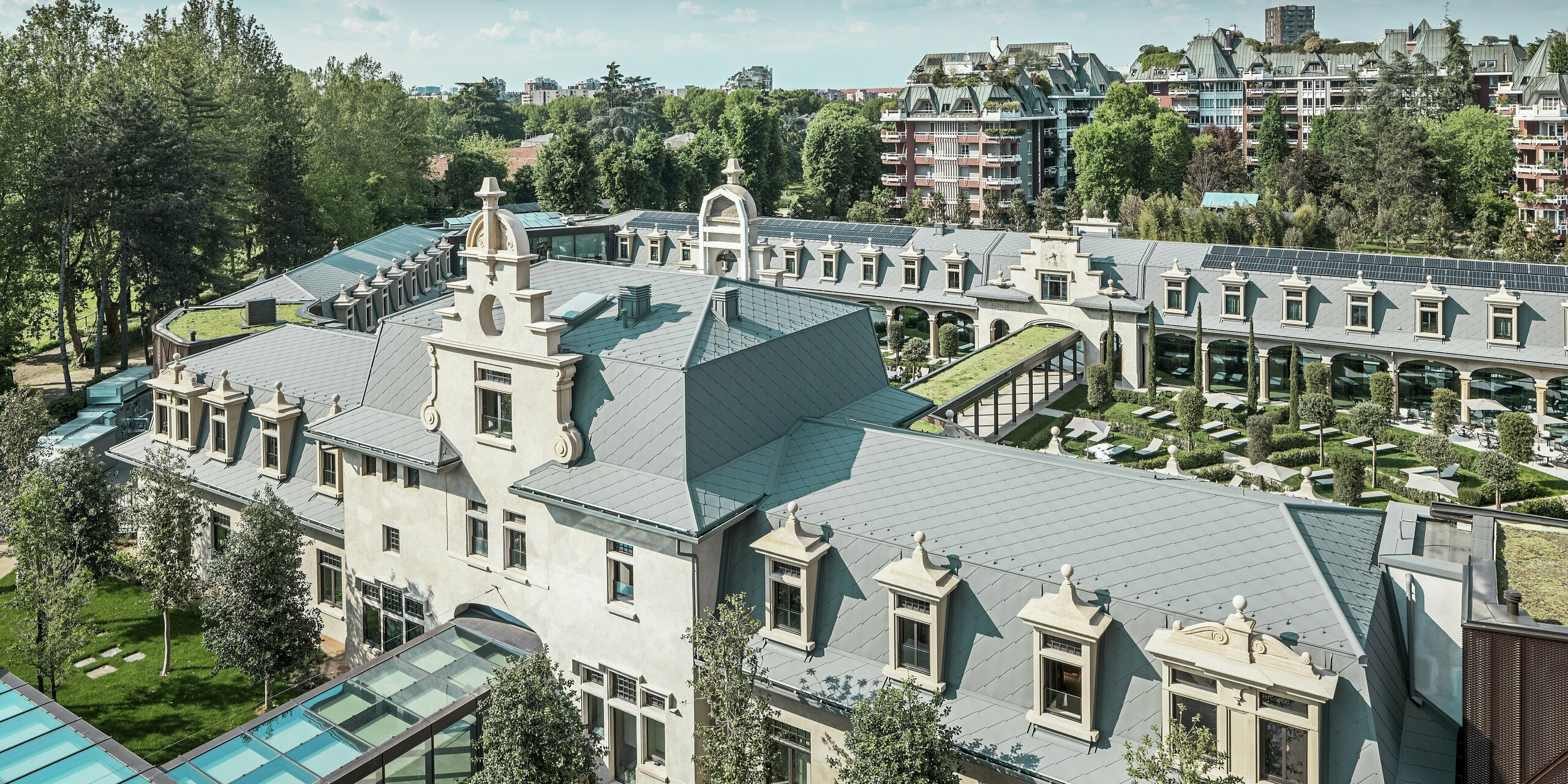 Celkový pohľad na impozantný komplex „De Montel – Terme Milano“ s pamiatkovo chránenou secesnou architektúrou a moderným wellness prostredím. Rozsiahla strešná krytina s PREFA strešnými šablónami 44 × 44 v odtieni P.10 svetlošedá dodáva komplexu elegantnú jednotnosť. Integrované fotovoltické moduly, zelené strechy a sklenené prepojenia medzi budovami odrážajú požiadavky na udržateľnosť a moderný dizajn v mestskom kontexte.