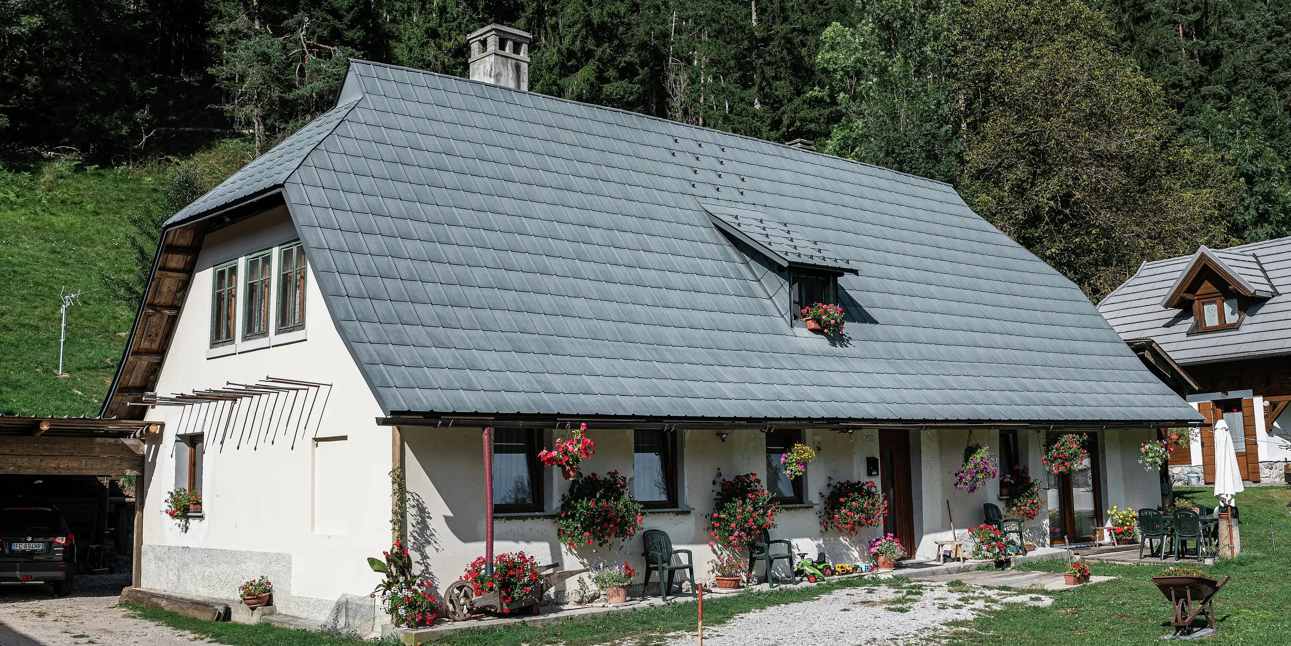 Idylický rodinný dom v Tarvisu v Taliansku je od roku 1972 pokrytý odolnými falcovanými škridlami PREFA. Rozsiahla, poveternostne odolná strecha z hliníka chráni dom už desaťročia pred alpskými poveternostnými podmienkami. Fasáda je zdobená množstvom kvetín, ktoré vytvárajú príjemnú atmosféru. Malá terasa s posedením láka k oddychu. V pozadí sa rozprestiera hustý les, ktorý harmonicky zapadá do prírody. Vľavo je vidieť krytý vjazd s zaparkovaným autom, zatiaľ čo vpravo obraz dotvára ďalší očarujúci drevený dom.