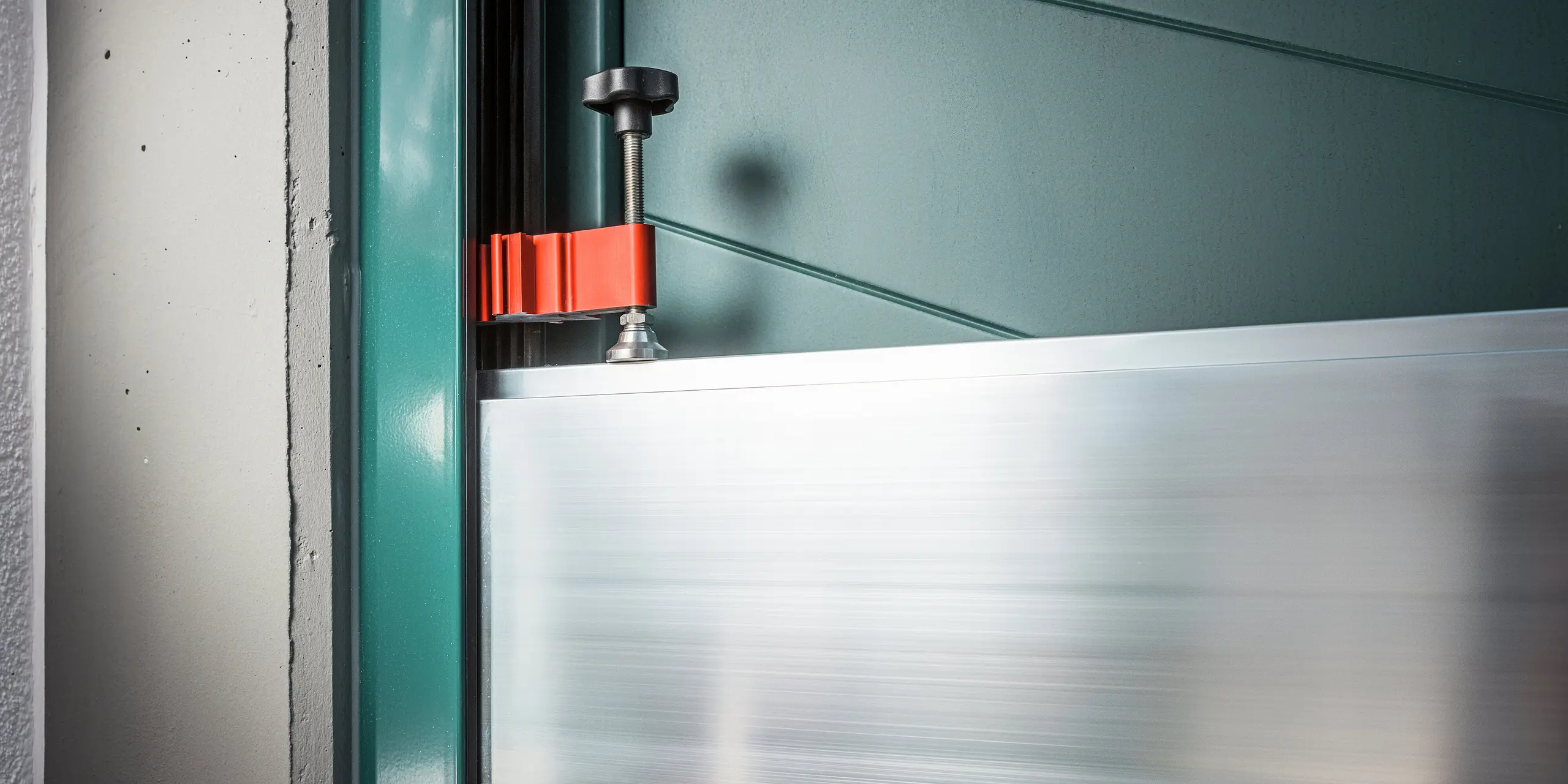 Detailný pohľad na upínací mechanizmus systému ochrany proti povodniam PREFA v oblasti dverí rodinného domu v Schwaz. Vidieť je presné upevnenie hliníkových hradidiel v bočných stenových profiloch pomocou červenej upínacej svorky a čiernej skrutkovej svorky. Vysoká kvalita spracovania a jednoduchá manipulácia podčiarkujú vhodnosť systému pre každodenné použitie – ideálny pre rýchlu ochranu proti povodniam pri vchodoch do domov.