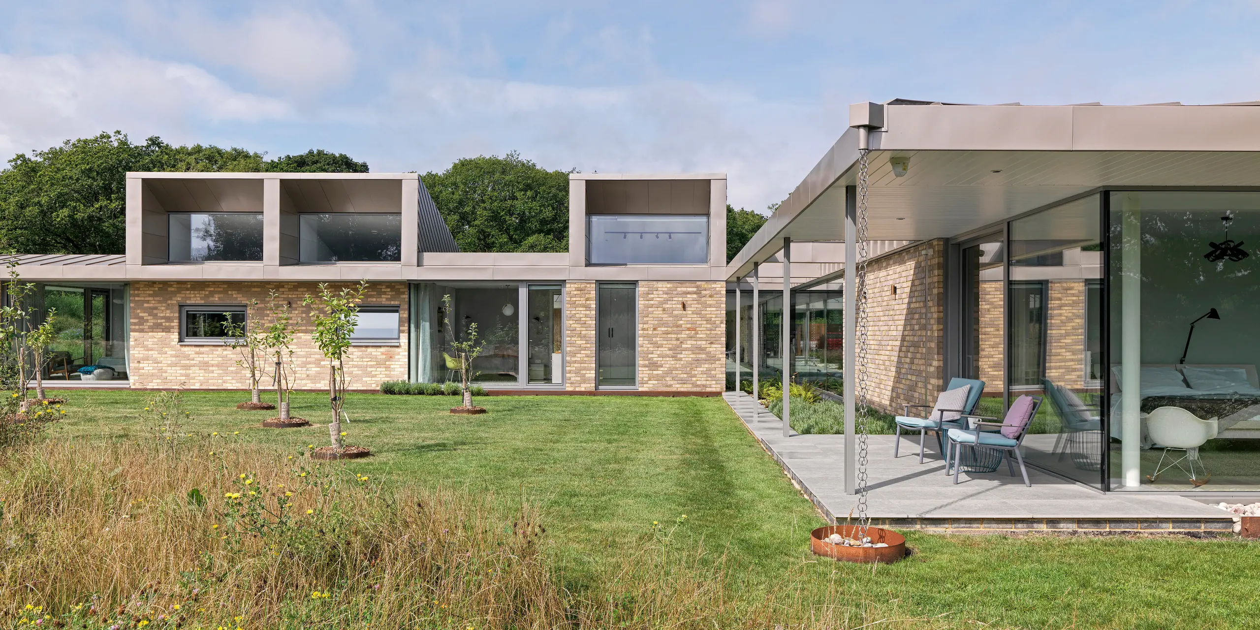 Bočný pohľad na luxusný obytný dom v West Sussex s výraznými nadstavbami na poschodí, obloženými PREFA fasádnymi šablónami 20 × 20 v P.10 bronzovej farbe. Plochá strecha je vyrobená z hliníka PREFALZ, tiež v odtieni P.10 bronzová, a podčiarkuje minimalistický dizajn. Rozsiahle sklenené fasády, krytá terasa s filigránovými podpierami a odkvapové reťaze dopĺňajú udržateľný architektonický koncept s dlhotrvajúcimi a bezúdržbovými hliníkovými produktmi PREFA.