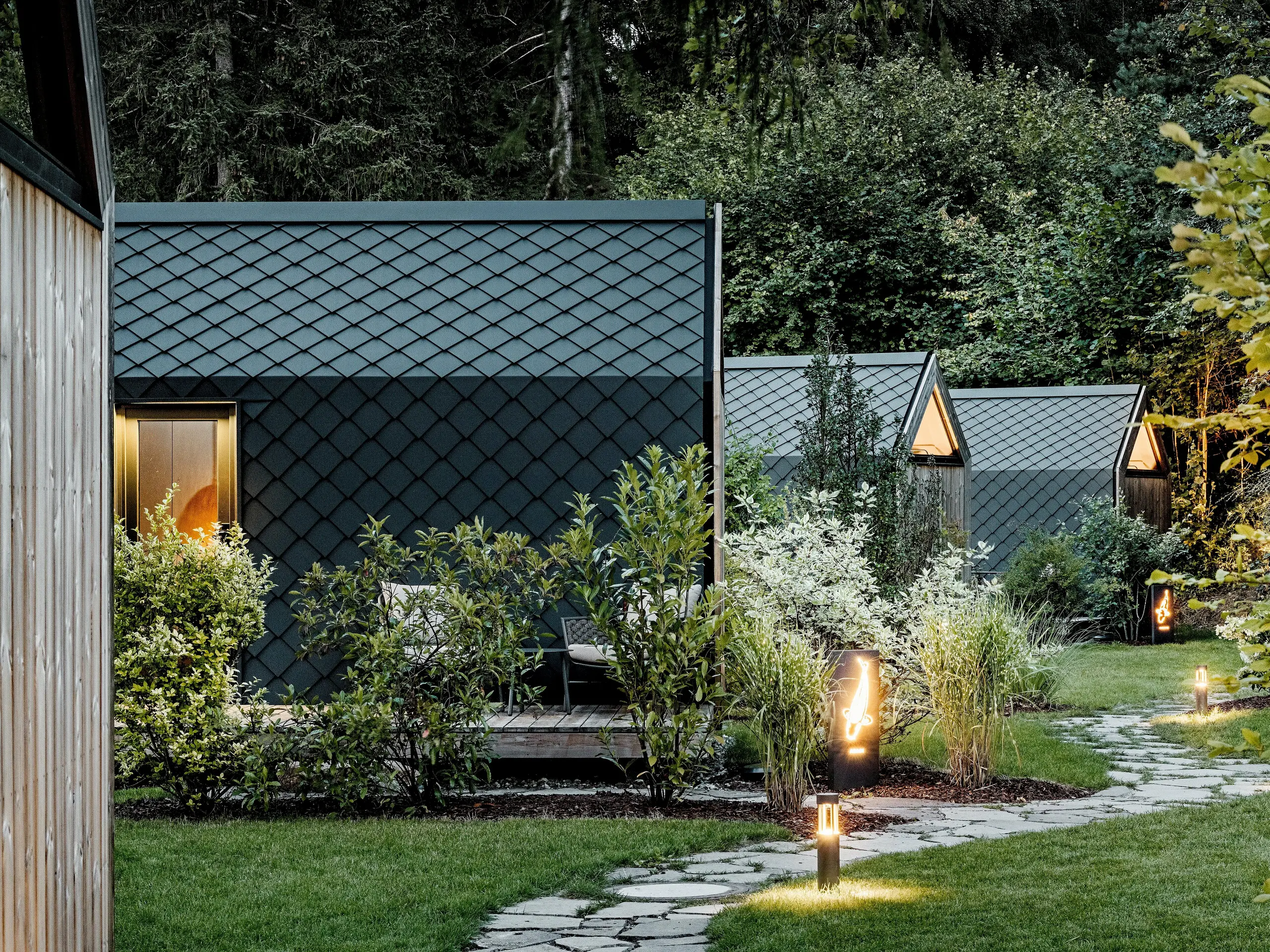 Osvetlená záhradná cestička vedie cez glampingový rezort Secret Koi Garden pri jazere Natterer See k viacerým minimalistickým minidomom. Tieto kompaktné stavby sú kompletne obložené hliníkovými strešnými a fasádnymi šablónami PREFA v odtieni P.10 antracit, ktorých štruktúra pripomínajúca rybiu kožu pôsobí vo večernom svetle elegantne. Uprostred bujnej zelene spájajú odolné a poveternostne odolné hliníkové povrchy PREFA kvalitnú architektúru s prírodou, pokojom a udržateľným dizajnom.