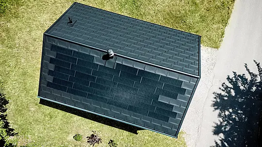 Pôsobivý záber z dronu zachytáva solárnu strechu PREFA domu Immi z vtáčej perspektívy. Čierne integrované solárne panely takmer neviditeľne prechádzajú do strešných panelov R.16 vo farbe P.10 čierna a vytvárajú súvislú strešnú plochu. Integrovaný hliníkový systém spája udržateľnú výrobu solárnej energie, vysokú odolnosť a čistú architektúru bez vizuálne rušivo pôsobiaceho fotovoltického systému namontovaného na povrchu strechy v alpskom prostredí.