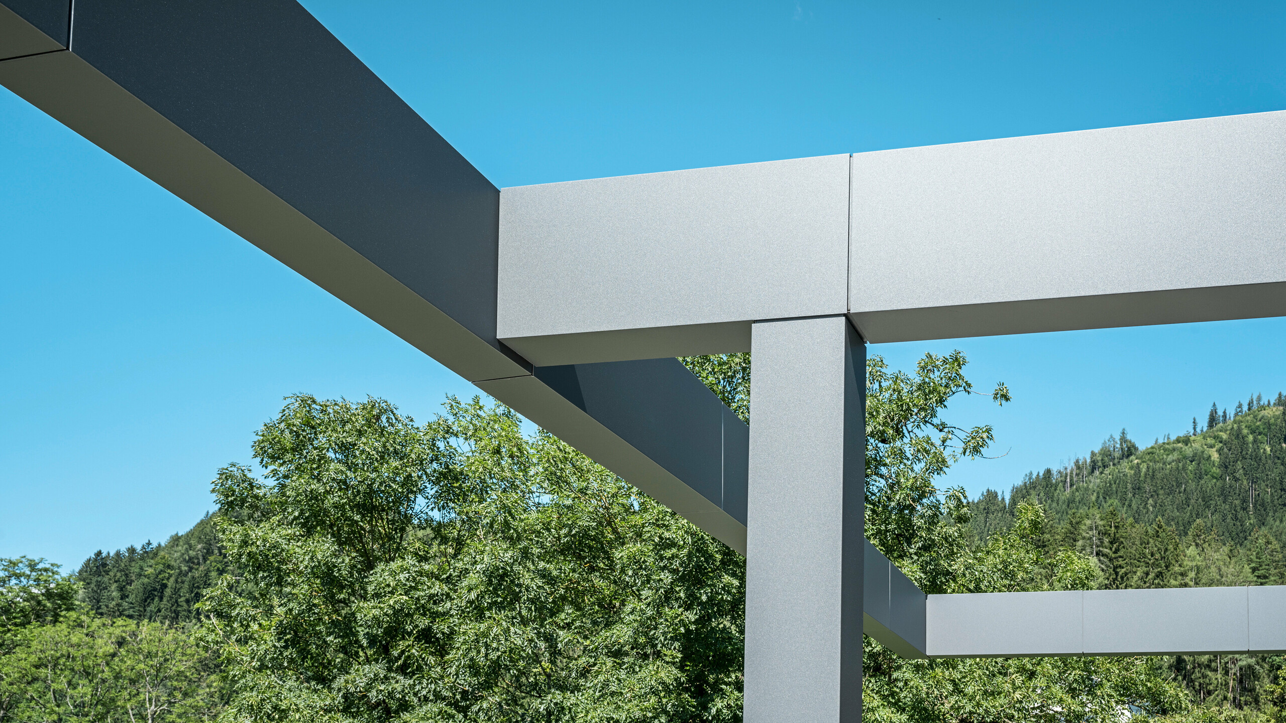 Moderné architektonické detaily terasovej strechy v rakúskom Pogieri, charakteristický PREFABOND v tmavošedej farbe. Čistá geometria vrchlíka s hranatými líniami a chladnou paletou farieb kontrastuje so živou zeleňou okolitých stromov a prírodnej krajiny. Táto konštrukcia je nielen funkčná, poskytuje tieň a prístrešie, ale je aj vizuálne príťažlivá, čo je dôkazom úspešnej kombinácie dizajnu a funkčnosti v modernej architektúre PREFA.