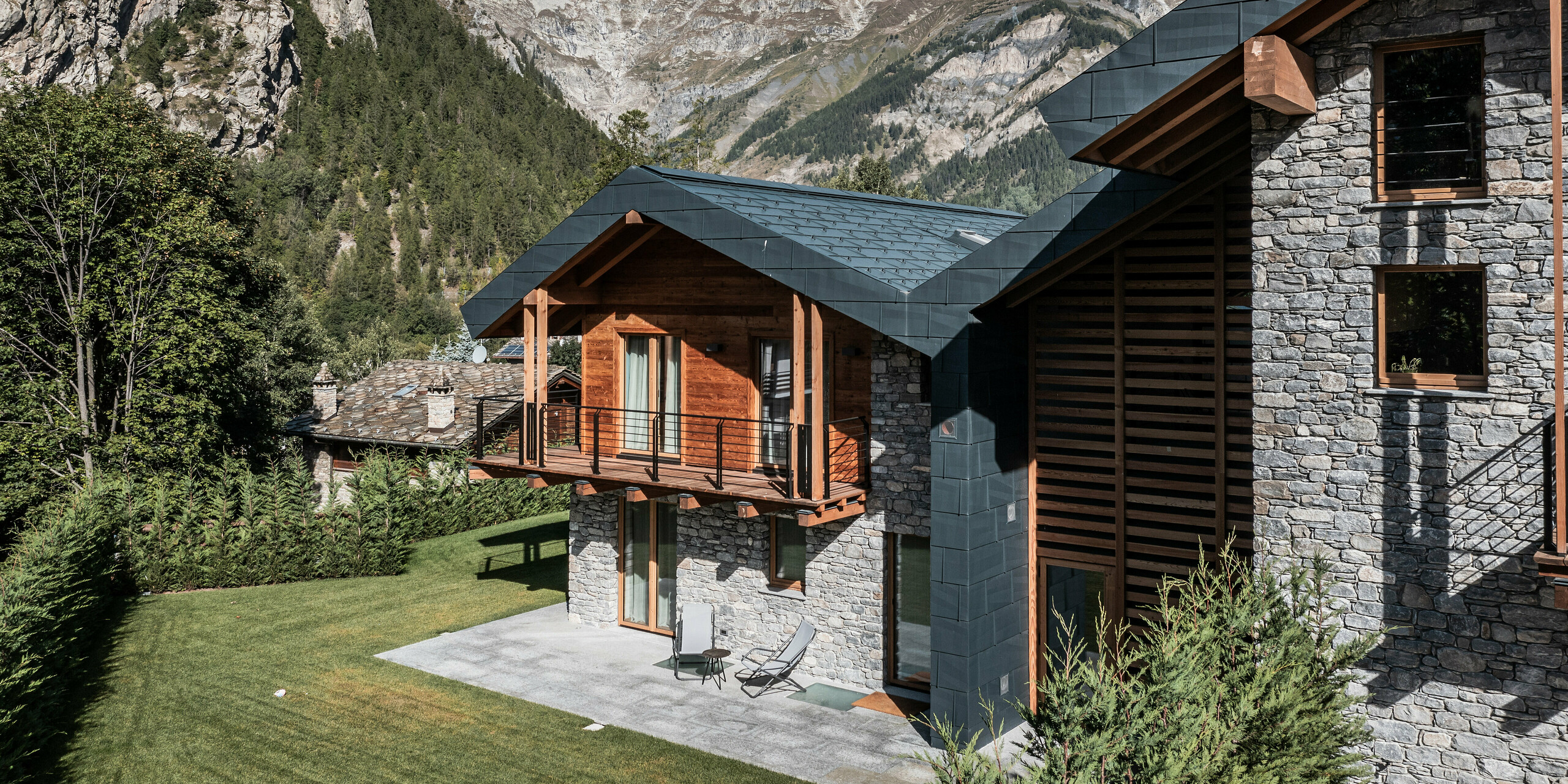 Nádherná vila v Courmayeur, zasadená do úchvatnej horskej krajiny údolia Aosta. Fasáda elegantne kombinuje drevo, kameň a PREFA hliník v kamennej šedej. Vila je charakteristická niekoľkými drevenými balkónmi a priestrannou terasou, ktorá pozýva k odpočinku a vychutnávaniu si nádherných výhľadov na okolité štíty. Svieža zeleň a historické kamenné domy v pozadí dotvárajú idylickú alpskú panorámu. Robustné a odolné strešné a fasádne panely FX.12 vyrobené z plechu PREFA poskytujú budove silnú ochranu pred zimnými podmienkami