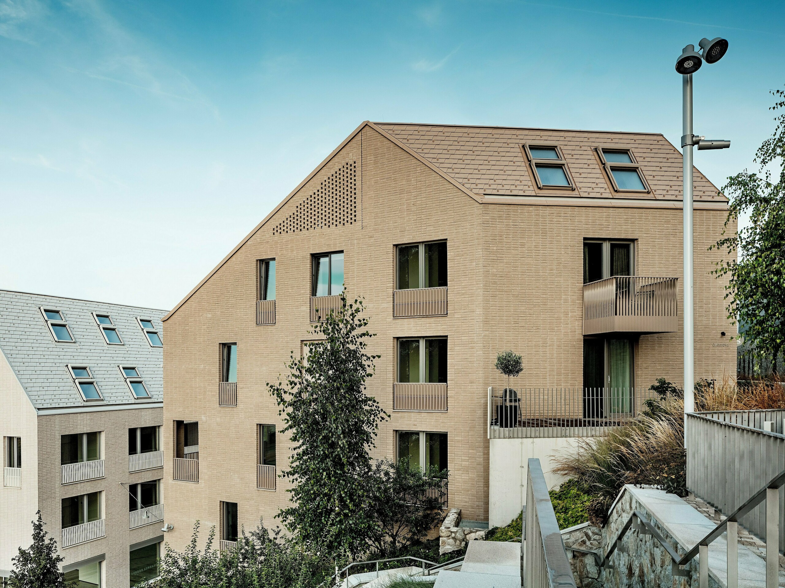 Moderné obytné budovy v mestskej štvrti Vydrica v Bratislave sa vyznačujú čistým, minimalistickým architektonickým výrazom a harmonickým zasadením do okolitého terénu. Strmé sedlové strechy s precízne uloženými hliníkovými strešnými šindľami PREFA vo farbách P.10 pieskovo hnedá a prefa biela vytvárajú vďaka štruktúrovanému povrchu a integrovaným strešným oknám výrazné vizuálne akcenty. Jednotné riešenie strechy a fasády prináša pokojný a esteticky hodnotný vzhľad. Hliník PREFA presviedča dlhou životnosťou, jednoduchým spracovaním, vysokou odolnosťou voči poveternostným vplyvom a umožňuje realizáciu náročných a udržateľných strešných riešení.