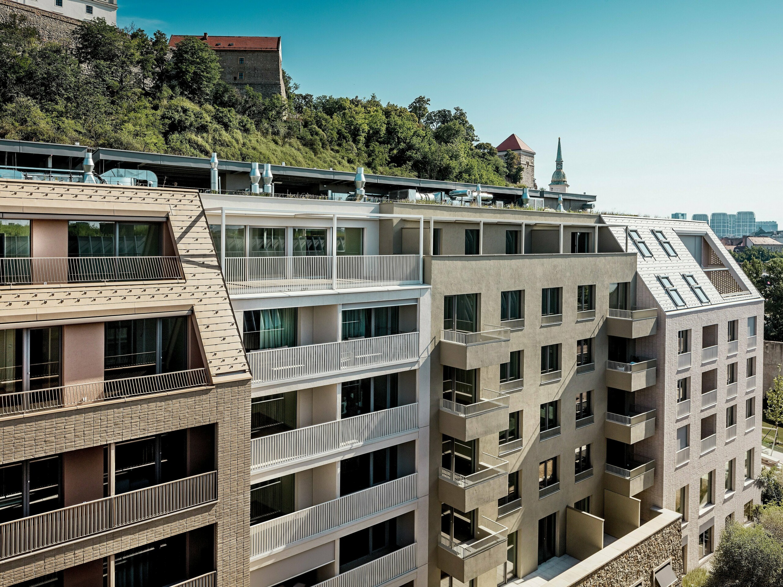 Mestský obytný komplex vo štvrti Vydrica v Bratislave prináša pôsobivé prepojenie modernej architektúry s historickou kulisou v blízkosti hradu. Strmé, precízne navrhnuté strechy s hliníkovými strešnými šindľami PREFA vo farbách pieskovo hnedá a prefa biela vytvárajú vďaka čistým líniám, vikierom a veľkorysým strešným terasám výrazné vizuálne akcenty. Kvalitné hliníkové riešenia umožňujú realizáciu komplexných tvarov striech a zároveň presviedčajú dlhou životnosťou, vysokou odolnosťou voči poveternostným vplyvom a minimálnymi nárokmi na údržbu, čím vytvárajú udržateľný a esteticky hodnotný obvodový plášť v mestskom prostredí.