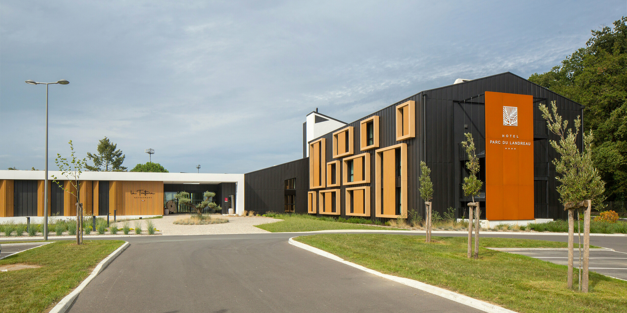 Prístup z parkoviska k hotelu Parc du Landreau v Les Herbiers, Francúzsko. Moderná budova je opláštená strešným a fasádnym systémom PREFALZ v čiernosivej farbe. Moderná architektúra je doplnená výraznými drevenými rámami okolo okien, ktoré harmonicky ladia s robustným hliníkovým opláštením. Priestranné predpolie, udržiavaná zeleň a nadčasový dizajn podčiarkujú príjemnú atmosféru hotela.