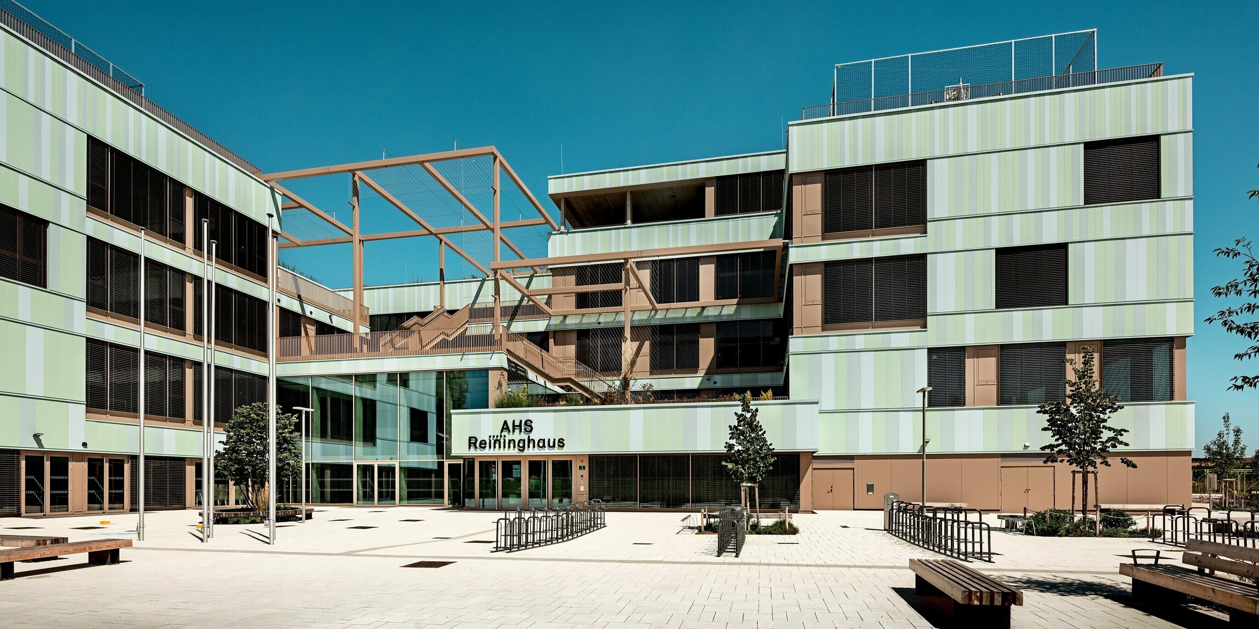 Das Bild zeigt den Eingangsbereich der AHS Reininghaus in Graz. Die Schule wurde mit PREFA-Sidings in Sonderfarben verkleidet. Die sanften Grüntöne und das Wechselspiel der vertikal angeordneten Aluminiumprofile verleihen dem Gebäude eine moderne und lebendige Ausstrahlung. Besonders hervorzuheben ist die klare, geometrische Struktur der Fassade, die durch die braunen Fensterrahmen und die modernen Jalousien ergänzt wird. Im Vordergrund ist der Eingangsbereich mit einer großzügigen, offenen Fläche zu sehen. Durch die Anordnung von Sitzbänken und Fahrradständern entsteht ein einladender Eindruck. Die PREFA Sidings sind nicht nur ästhetisch ansprechend, sondern auch äußerst widerstandsfähig gegenüber Witterungseinflüssen und äußerst pflegeleicht. Die Fassade trägt zur Energieeffizienz des Gebäudes bei, während das gesamte Design die Funktionalität der Bildungseinrichtung unterstreicht und sie perfekt in die Umgebung integriert. Der offene, moderne Stil und die nachhaltige Materialwahl machen das Gebäude zu einem herausragenden Beispiel für zeitgenössische Architektur.