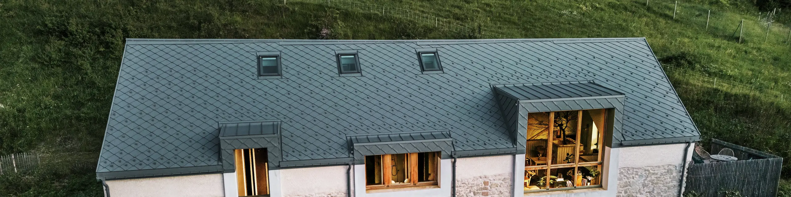 Večerný pohľad na obnovenú stodolu „La Grange à Gaby“ v Engins, Francúzsko. Budova kombinuje tradičné kamenné murivo s modernou hliníkovou strechou PREFA zo šablón s rozmermi 29 × 29 cm. Veľké okná a osvetlený interiér podčiarkujú prívetivú atmosféru nového kultúrneho centra.