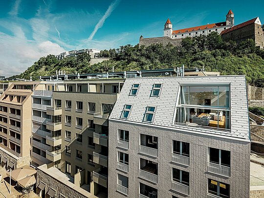 Moderné riešenie striech v mestskej štvrti Vydrica v Bratislave s výhľadom na hradný areál: Strmo sklonené strešné plochy s veľkorysými okennými otvormi a čistými líniami elegantne dotvárajú hliníkové strešné šindle PREFA vo farbách prefa biela a pieskovo hnedá. Precízne spracovanie a harmonické prepojenie strechy s fasádou vytvárajú výrazný vizuálny prvok nad strechami mesta. Hliník PREFA presviedča nízkou hmotnosťou, vysokou odolnosťou voči poveternostným vplyvom a dlhou životnosťou – ideálne riešenie pre komplexné a architektonicky náročné obytné stavby.