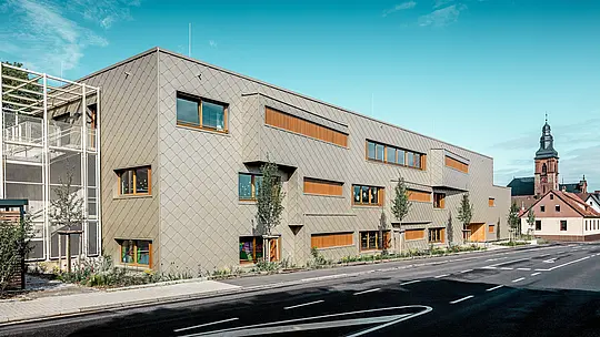 V jasnom dennom svetle sa moderná materská škola v Alzenau s výraznou hliníkovou fasádou PREFA z bronzových šablón 44 × 44 ukazuje v plnej kráse. Rovnomerná šupinová štruktúra, prerušovaná posunutými oknami a vertikálnymi drevenými lamelami, dodáva fasáde hĺbku a rytmus. Vľavo na obrázku je viditeľné filigránové schodisko s kovovou mriežkovou konštrukciou, ktoré zaisťuje transparentnosť a ľahkosť. V pozadí stojí neskorogotický kostol v Alzenau – symbol harmonického spojenia historickej stavebnej kultúry a modernej architektúry. Bezúdržbový, odolný fasádny systém PREFA podporuje udržateľný, funkčný a esteticky vysoko kvalitný dizajn tejto budovy vzdelávacieho zariadenia orientovaného na budúcnosť.