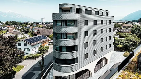 Letecký pohľad na Bernerhaus v Buchsi s výraznou trojuholníkovou kubatúrou a jemne zaoblenými balkónmi. Fasáda z PREFA šindľov v odtieni P.10 svetlošedá obklopuje budovu ako šupinatá hliníková koža a vytvára živú hru svetla a tieňa. Nad masívnym betónovým podstavcom s elegantnými arkádami pôsobí ľahká, odolná proti poveternostným vplyvom PREFA hliníková fasáda mimoriadne precízne, trvalo a s nízkymi nárokmi na údržbu – moderný pohľad upútavajúci uprostred alpského mestského prostredia.