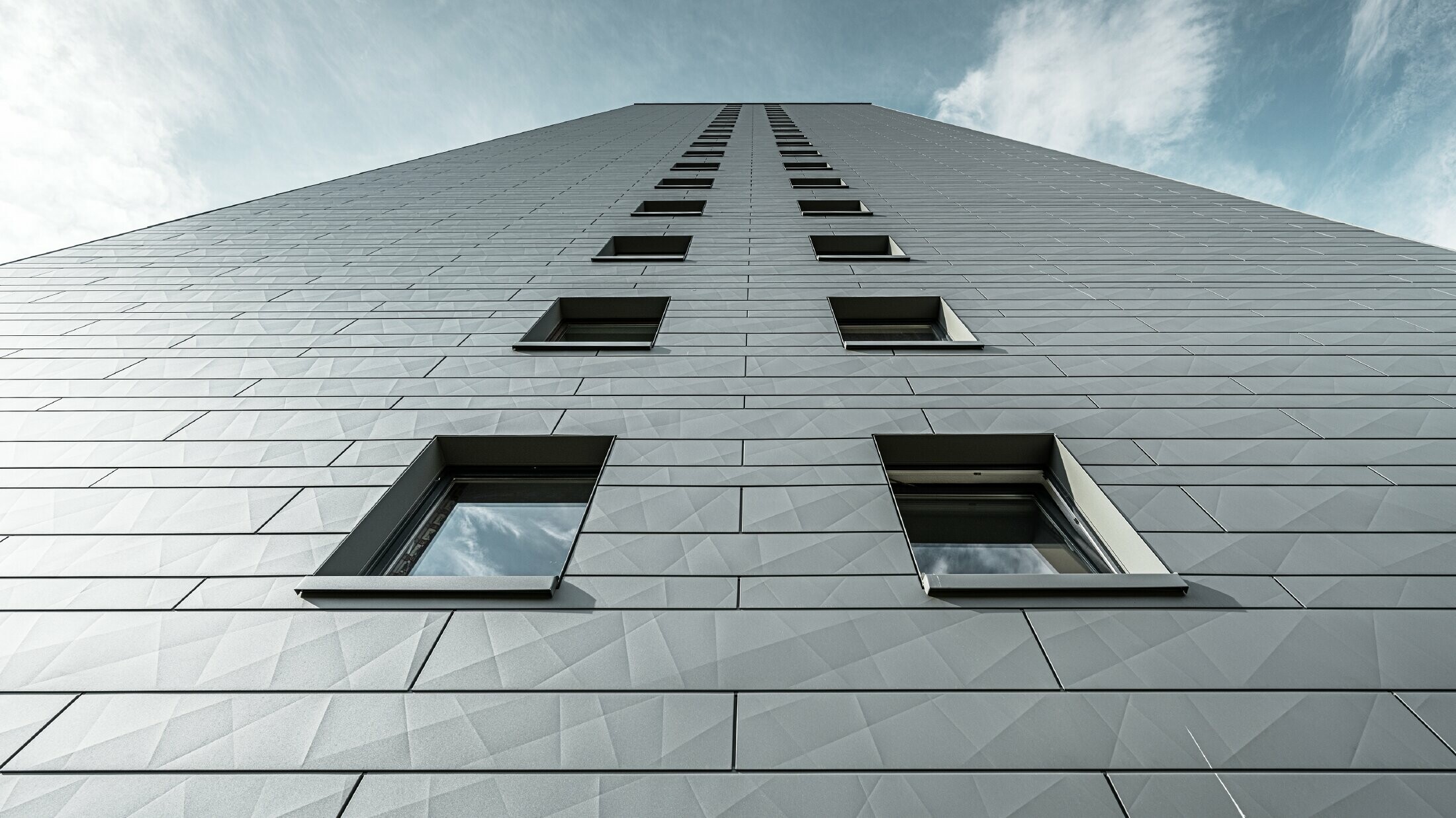 Blick an der Fassade hoch an einem vielstöckigen Hochhaus, die Fassade ist verkleidet mit waagrechten Siding.X Fassadenpaneelen von PREFA in Hellgrau.