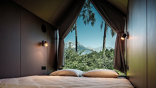 Pohľad z postele minimalistického minidomu v Secret Koi Garden pri jazere Natterer See: Vďaka veľkej trojuholníkovej sklenenej fasáde sa priestor otvára k prírode so stromami a výhľadom na tirolskú horskú krajinu. Minimalistický interiér smeruje pozornosť von a podčiarkuje koncept spomalenia. Architektúru chránia odolné hliníkové rímsy PREFA, ktoré spájajú dizajn, odolnosť voči poveternostným vplyvom a udržateľnosť.