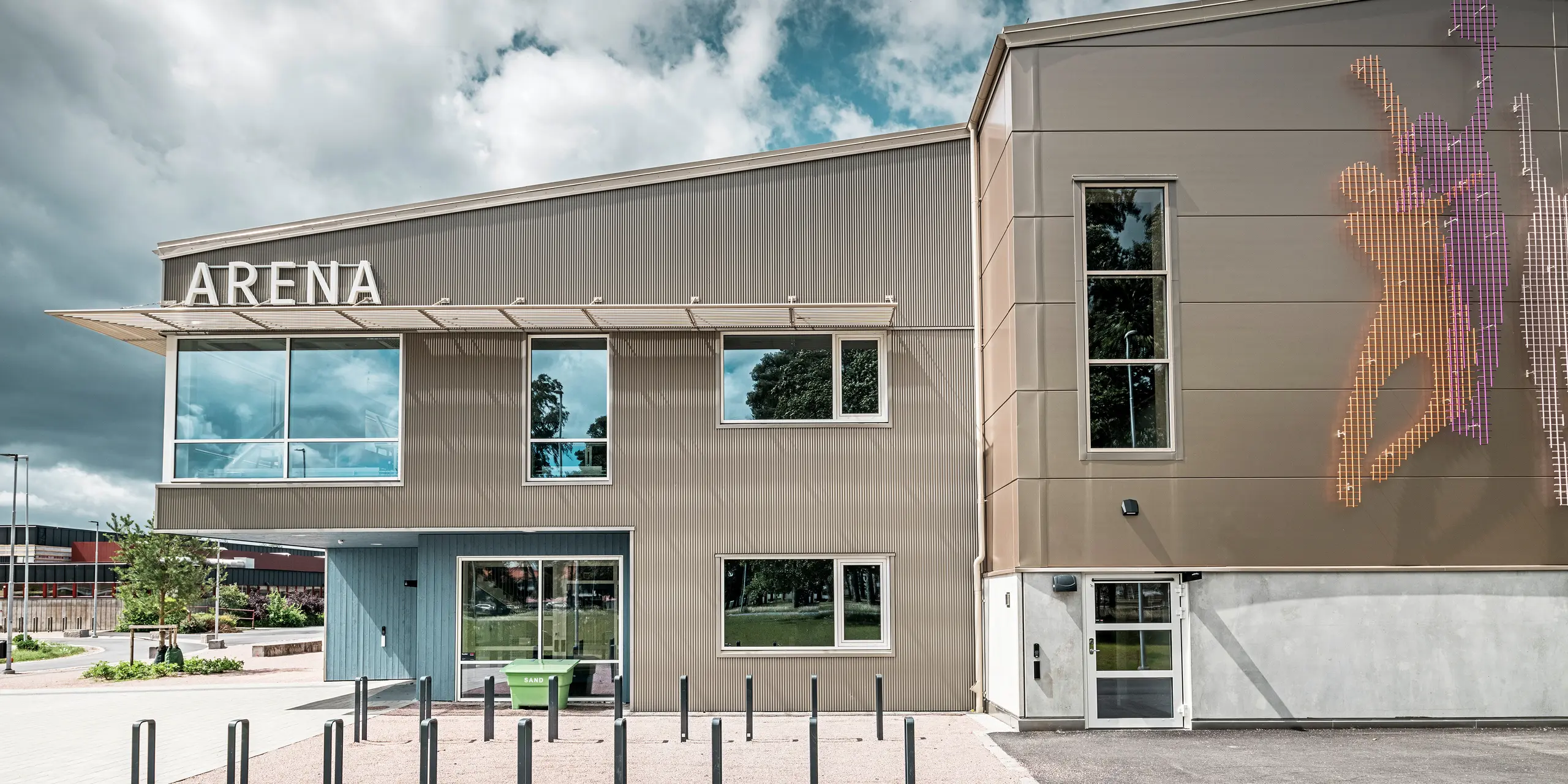 Bočný pohľad na vstupný priestor do Park Areny vo švédskom meste Värnamo s modernou fasádou PREFA z bronzového prelamovaného profilu. Hliníková fasáda je odolná a vytvára pôsobivé efekty svetla a tieňa. V pravej časti obrázka je umelecky navrhnutá nástenná maľba zobrazujúca športové postavy, ktorá podčiarkuje dynamickú atmosféru športovej haly. Veľké okná prepúšťajú do budovy dostatok svetla a zdôrazňujú moderný dizajn.