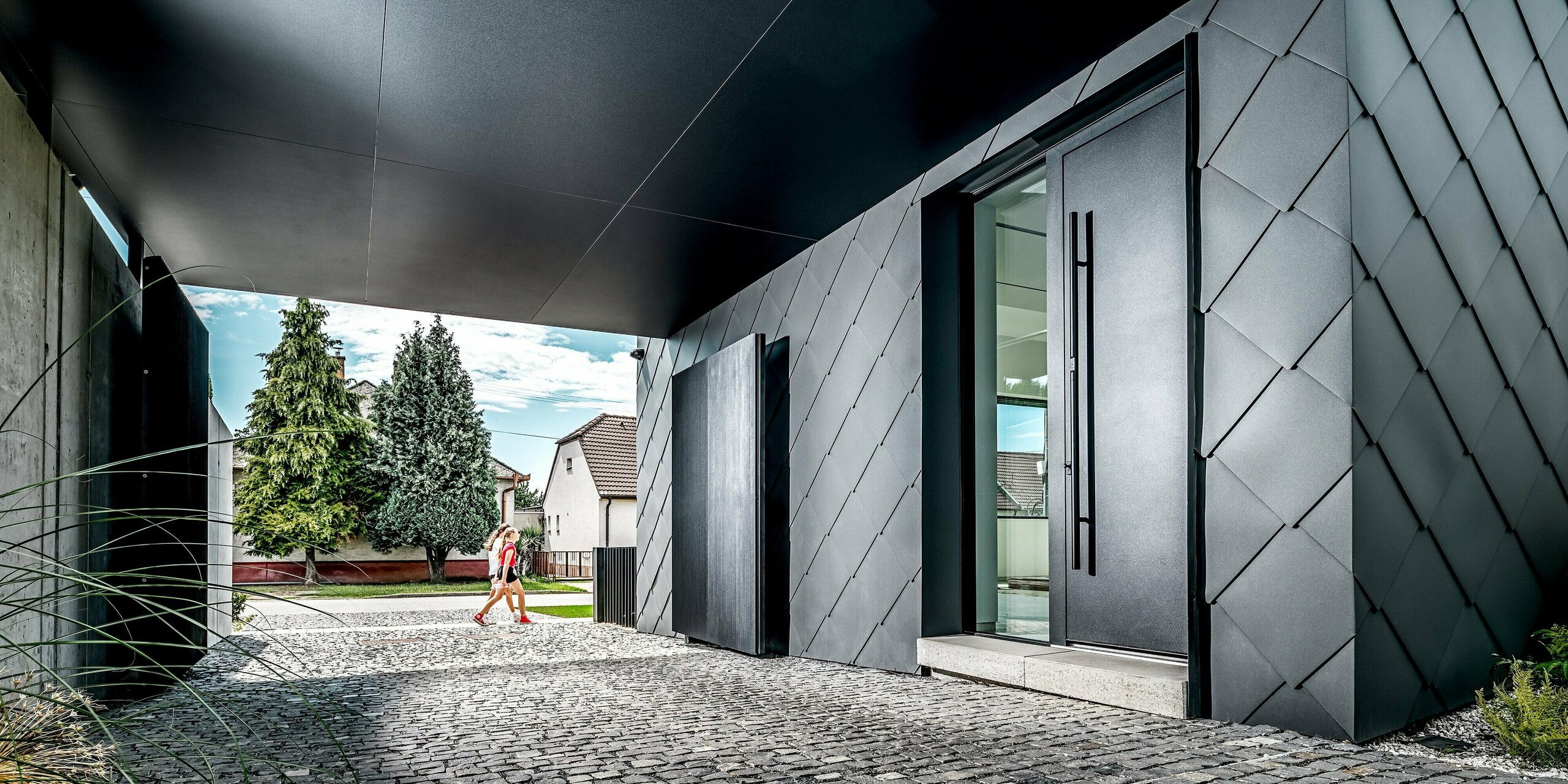 Tento obrázok zachytáva vstupný a priechodný priestor moderného rodinného domu v Trnave, ktorého výrazná fasáda je kompletne obložená fasádnymi šablónami PREFA 44 × 44 v odtieni P.10 tmavošedá. Presná kosoštvorcová štruktúra dodáva budove elegantný, futuristický vzhľad a zdôrazňuje čisté línie architektúry. Odolné hliníkové produkty zaisťujú trvalú ochranu pred poveternostnými vplyvmi a kvalitný, dlhotrvajúci vzhľad. V pozadí vytvára zeleň obytného prostredia prirodzený kontrast k tmavému plášťu budovy.