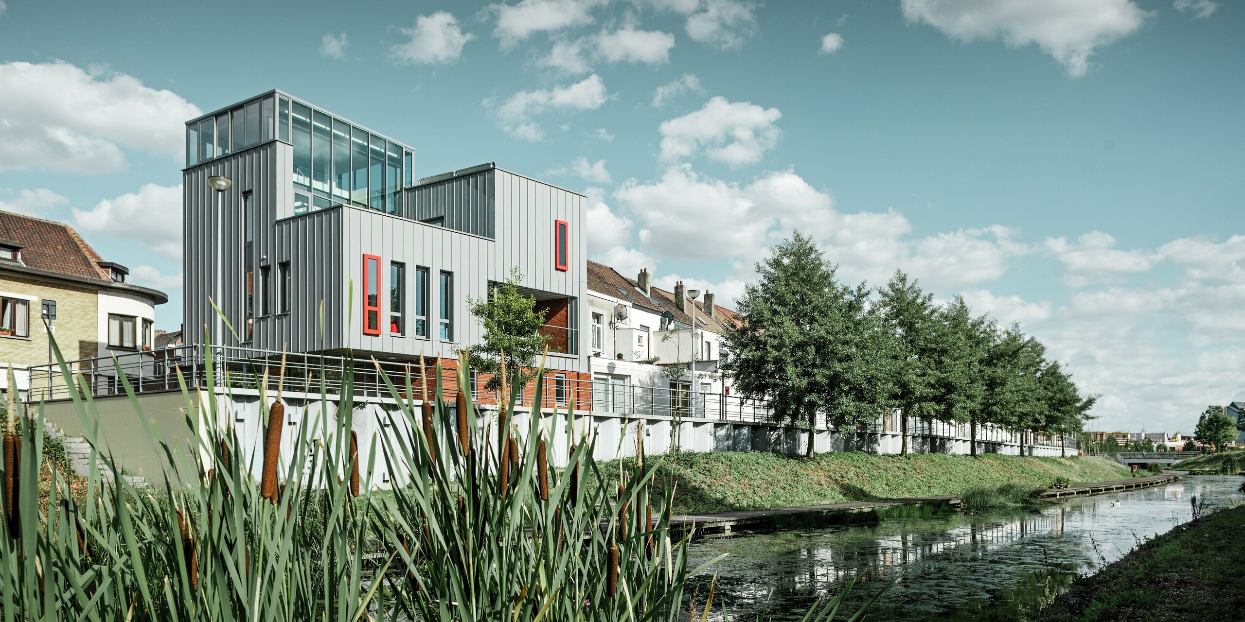 Architektonický skvost pri vode: Moderná obytná a administratívna budova zaujme hliníkovou fasádou PREFALZ v patinovo šedej farbe s vertikálnou stojatou falcovou štruktúrou. Farebne odlíšené okná v červenej farbe vytvárajú cielené akcenty. Kombinácia skla, hliníka a červenej tehly vytvára harmonický celkový obraz – odolný voči poveternostným vplyvom, ľahko udržiavateľný a trvácny.