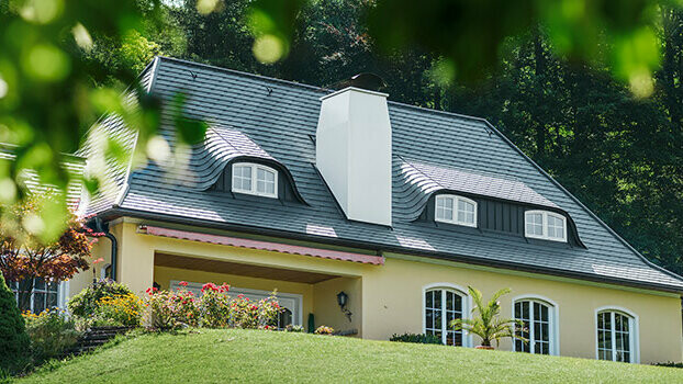 Rodinný dom s čerstvo zrekonštruovanou strechou s použitím PREFA falcovaných šindľov antracitovej farby, s oblúkovými vikiermi a bielym komínom.