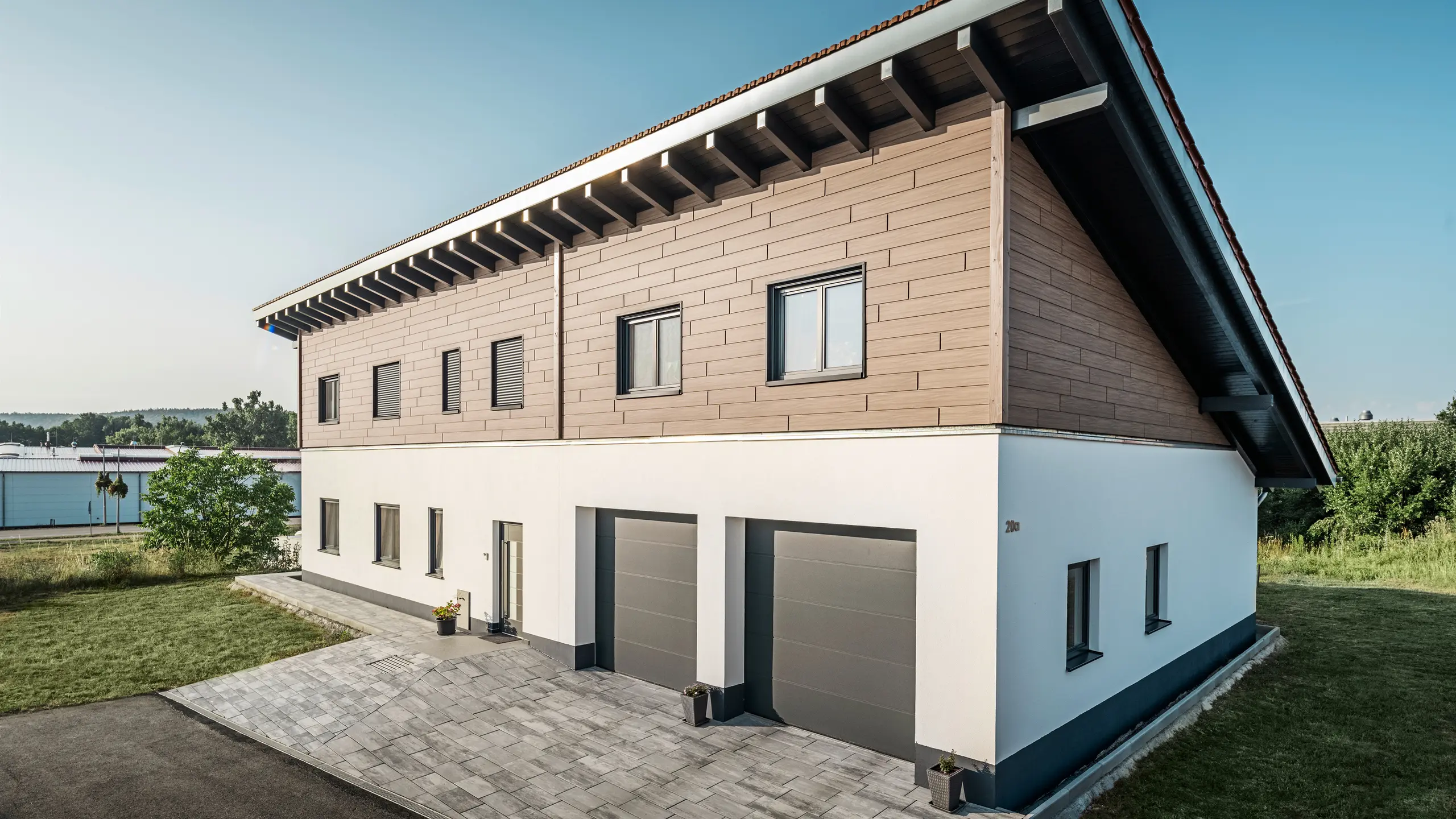 Pohľad na rodinný dom s jednoplášťovou strechou v Brucku. Horné podlažie je obložené obkladom PREFA Siding v vlašský orech hnedý farbe. Na pravej strane obrázka sú viditeľné dve garážové brány v antracitovej farbe. Vstupné dvere sa nachádzajú v strede budovy. Sokel je v jednoduchej bielej farbe. 