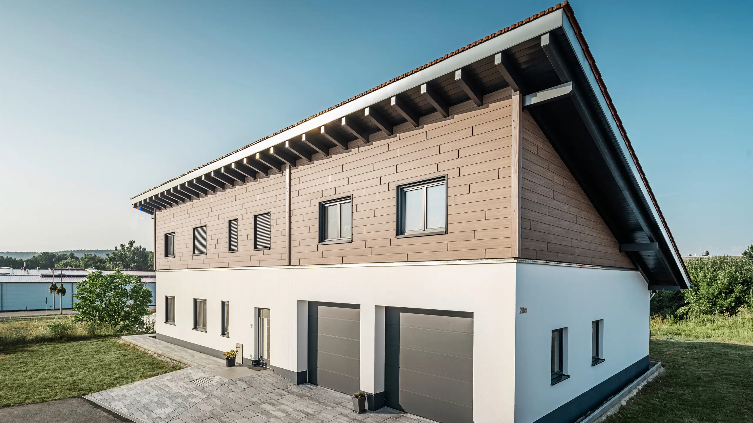 Pohľad na rodinný dom s jednoplášťovou strechou v Brucku. Horné podlažie je obložené obkladom PREFA Siding v vlašský orech hnedý farbe. Na pravej strane obrázka sú viditeľné dve garážové brány v antracitovej farbe. Vstupné dvere sa nachádzajú v strede budovy. Sokel je v jednoduchej bielej farbe. 