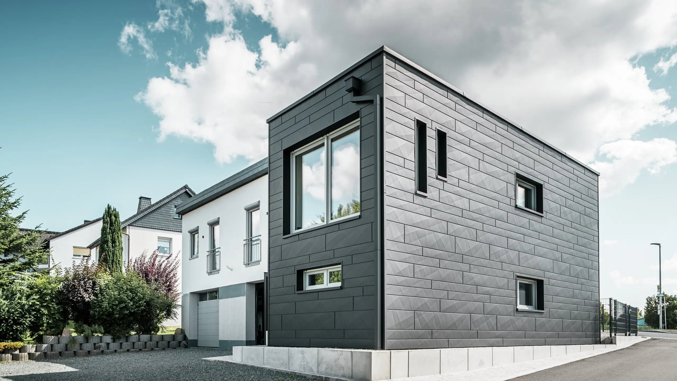 Štýlová a moderná fasáda zrekonštruovaného rodinného domu v Teuschnitzi je tvorená jasne štruktúrovanými panelmi PREFA Siding.X v čiernej farbe P.10. Geometrické usporiadanie kvalitných plechových panelov dodáva budove výraznú, nadčasovú estetiku. Okná sú orámované čiernymi hliníkovými kompozitnými doskami, čo harmonicky dotvára dizajn. Integrovaný odvodňovací systém PREFA s hranatými zvodmi poskytuje funkčné a estetické riešenie.  Translated with DeepL.com (free version)