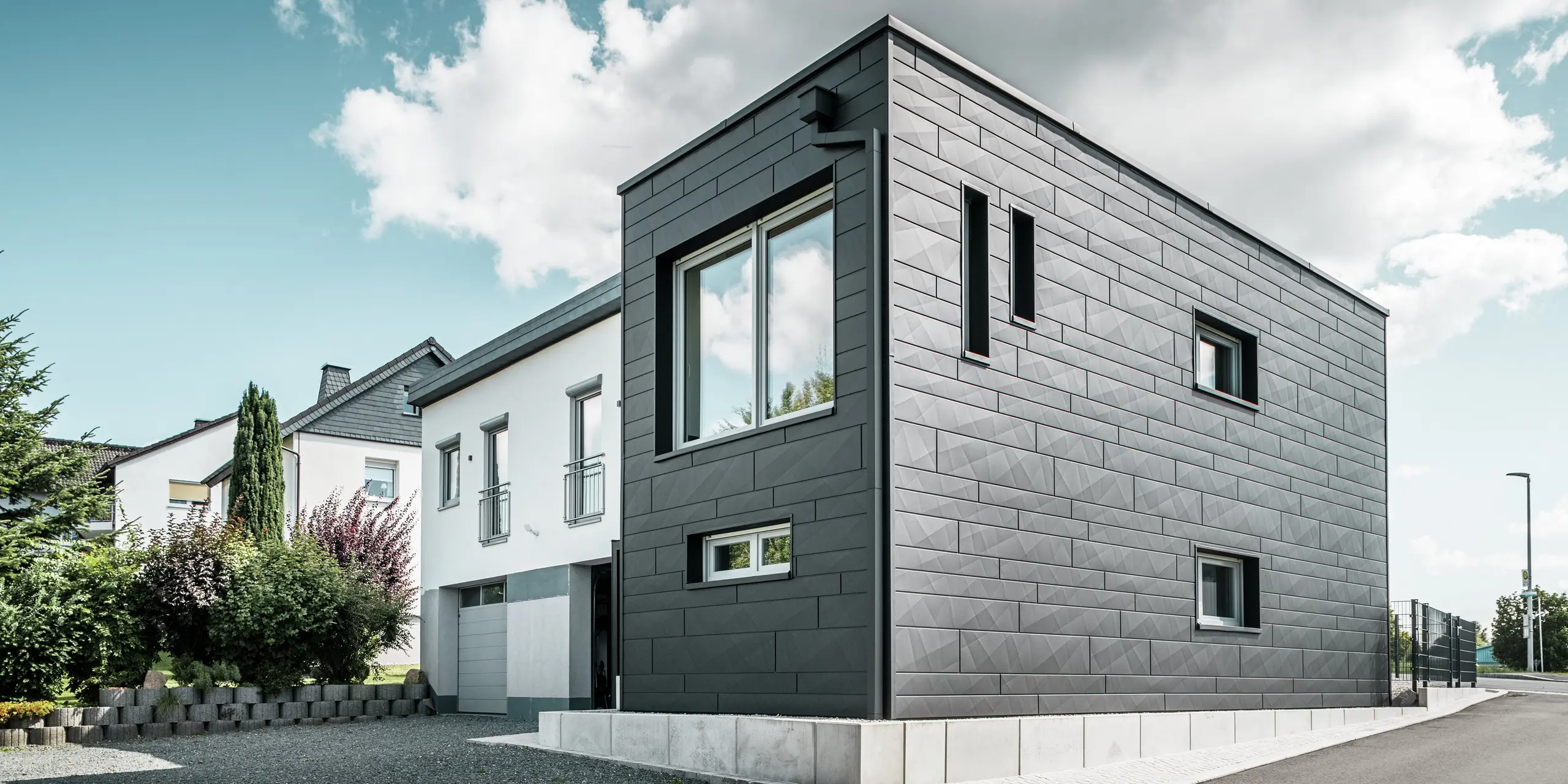 Štýlová a moderná fasáda zrekonštruovaného rodinného domu v Teuschnitzi je tvorená jasne štruktúrovanými panelmi PREFA Siding.X v čiernej farbe P.10. Geometrické usporiadanie kvalitných plechových panelov dodáva budove výraznú, nadčasovú estetiku. Okná sú orámované čiernymi hliníkovými kompozitnými doskami, čo harmonicky dotvára dizajn. Integrovaný odvodňovací systém PREFA s hranatými zvodmi poskytuje funkčné a estetické riešenie.  Translated with DeepL.com (free version)