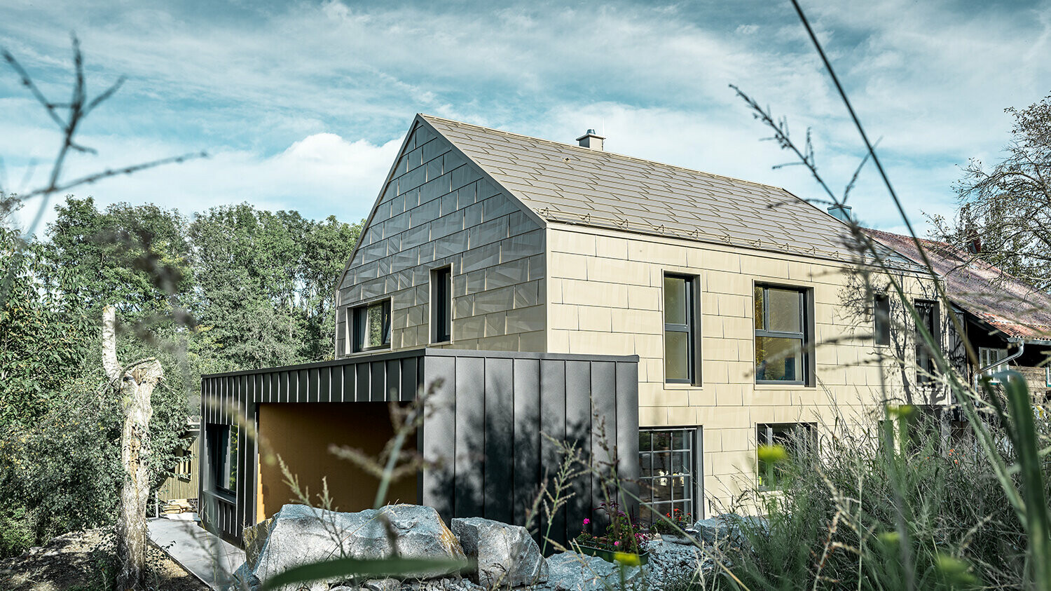 Obytný dom bez previsnutej strechy je na streche a na fasáde obložený hliníkovým panelom FX.12 od firmy PREFA vo farbe bronzová. Hneď vedľa stojí garáž s plochou strechou s fasádou s uhlovou stojatou drážkou.