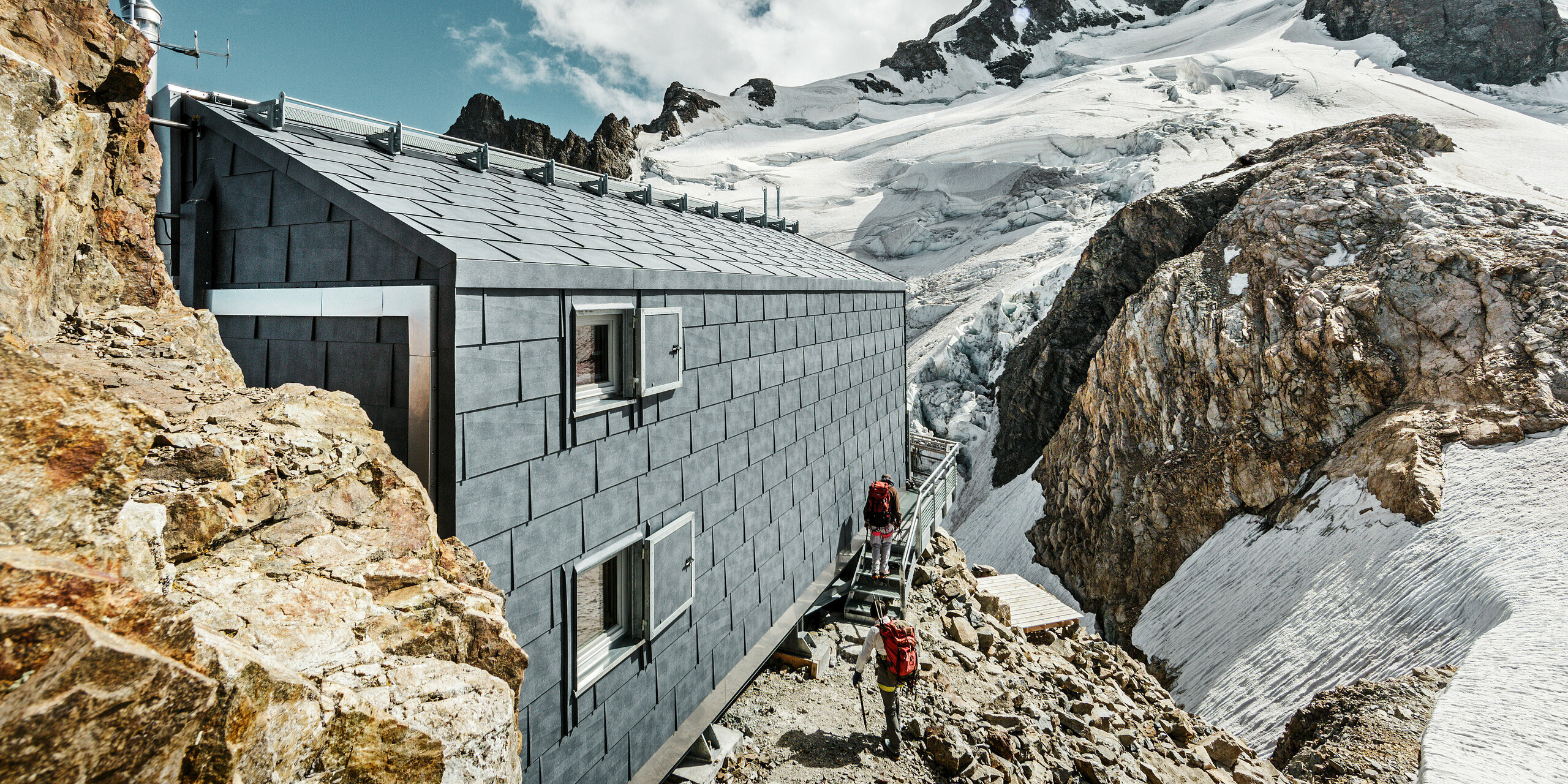 Seitliche Ansicht der Refuge de l'Aigle Berghütte mit steingrauen FX.12 Dach- und Fassadenpaneelen von PREFA, eingebettet in eine beeindruckende Gletscherlandschaft.