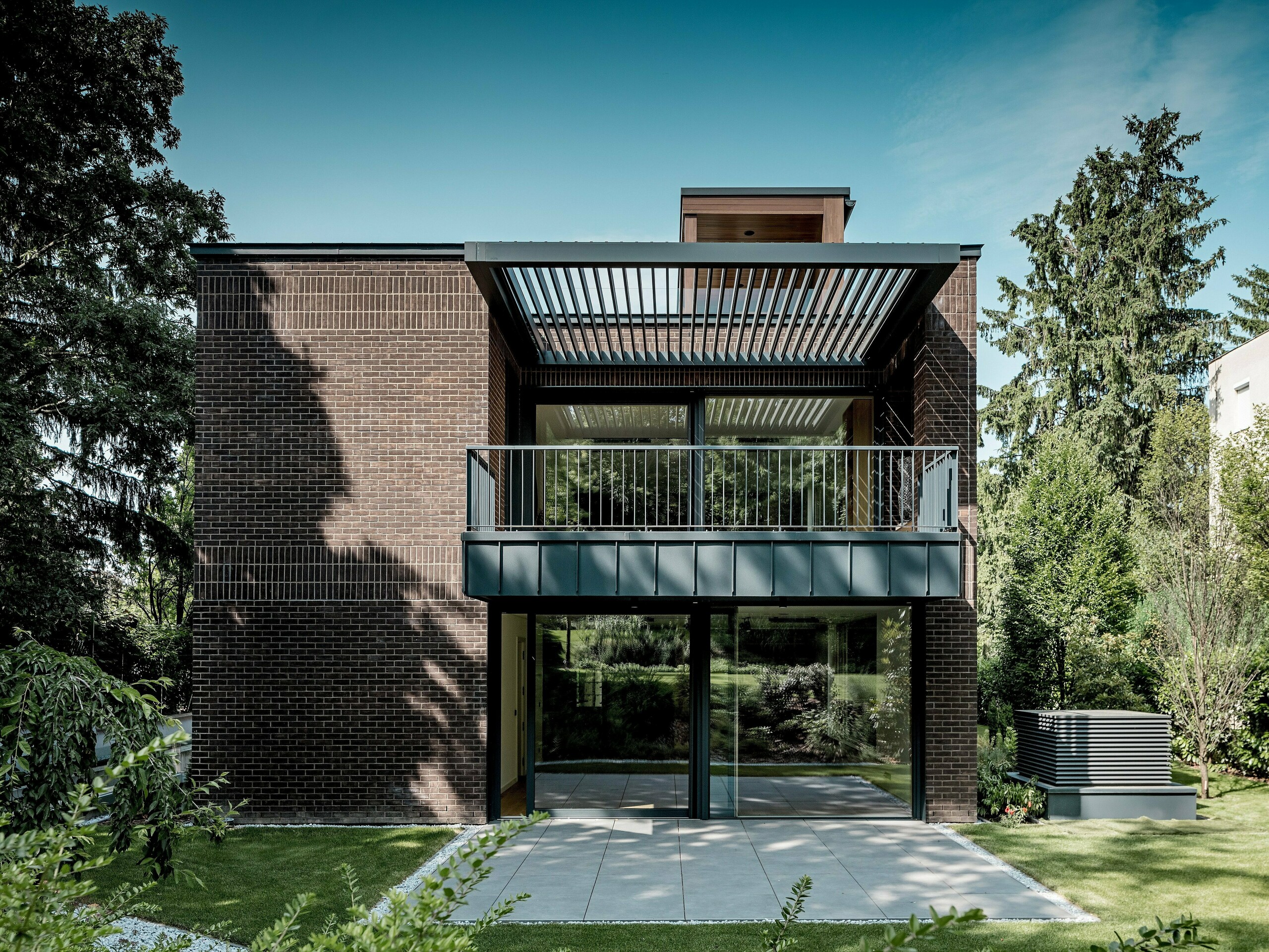 Záber zachytáva záhradnú stranu modernej viacgeneračnej vily v Budapešti s jasne štruktúrovanou fasádou z tmavočerveného klinkeru a hliníka PREFALZ v odtieni P.10 antracit. Veľký balkón s jemnou kovovou konštrukciou a lamelami nad ním vytvára zaujímavú hru svetla a tieňa. Presklené plochy od podlahy po strop otvárajú interiér smerom k zelenej záhrade. Precízne vyhotovené hliníkové detaily zdôrazňujú dlhú životnosť, odolnosť voči poveternostným vplyvom a architektonickú čistotu riešení PREFA, ktoré dodávajú budove výrazný a nadčasový charakter.