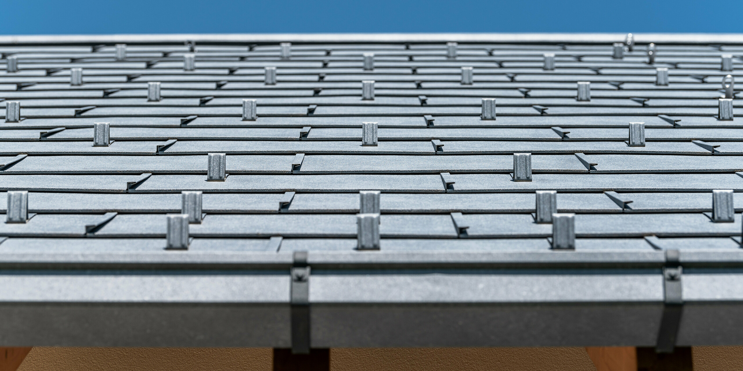 Detailný pohľad na strechu PREFA pokrytú strešným panelom R.16 vo farbe P.10 antracit. Na obrázku sú precízne spracované hliníkové strešné panely. Záber vyzdvihuje nápadnú štruktúru a moderný dizajn strechy, ktorý kontrastuje s jasne modrou oblohou a podčiarkuje inovatívne a odolné riešenie zastrešenia PREFA.