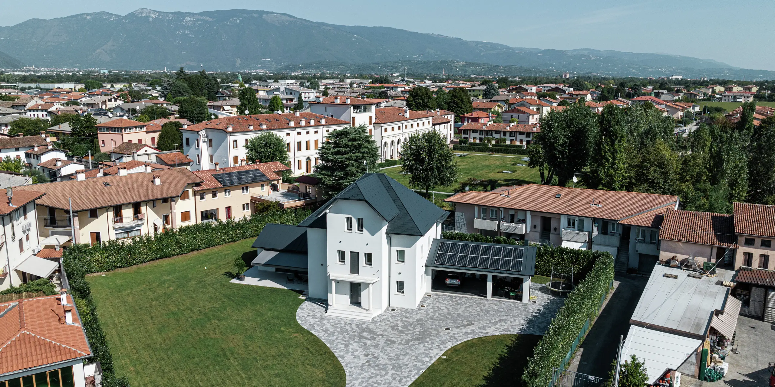 Letecký pohľad na moderný rodinný dom v Marano Vicentino v Taliansku s robustnou strechou z PREFA strešných šindľov v antracitovej farbe P.10. Priľahlé vedľajšie budovy sú pokryté klasickou dvojitou stojatou falcovou krytinou z hliníka. Vysoko kvalitná hliníková krytina zaisťuje dlhotrvajúci a estetický vzhľad strechy.