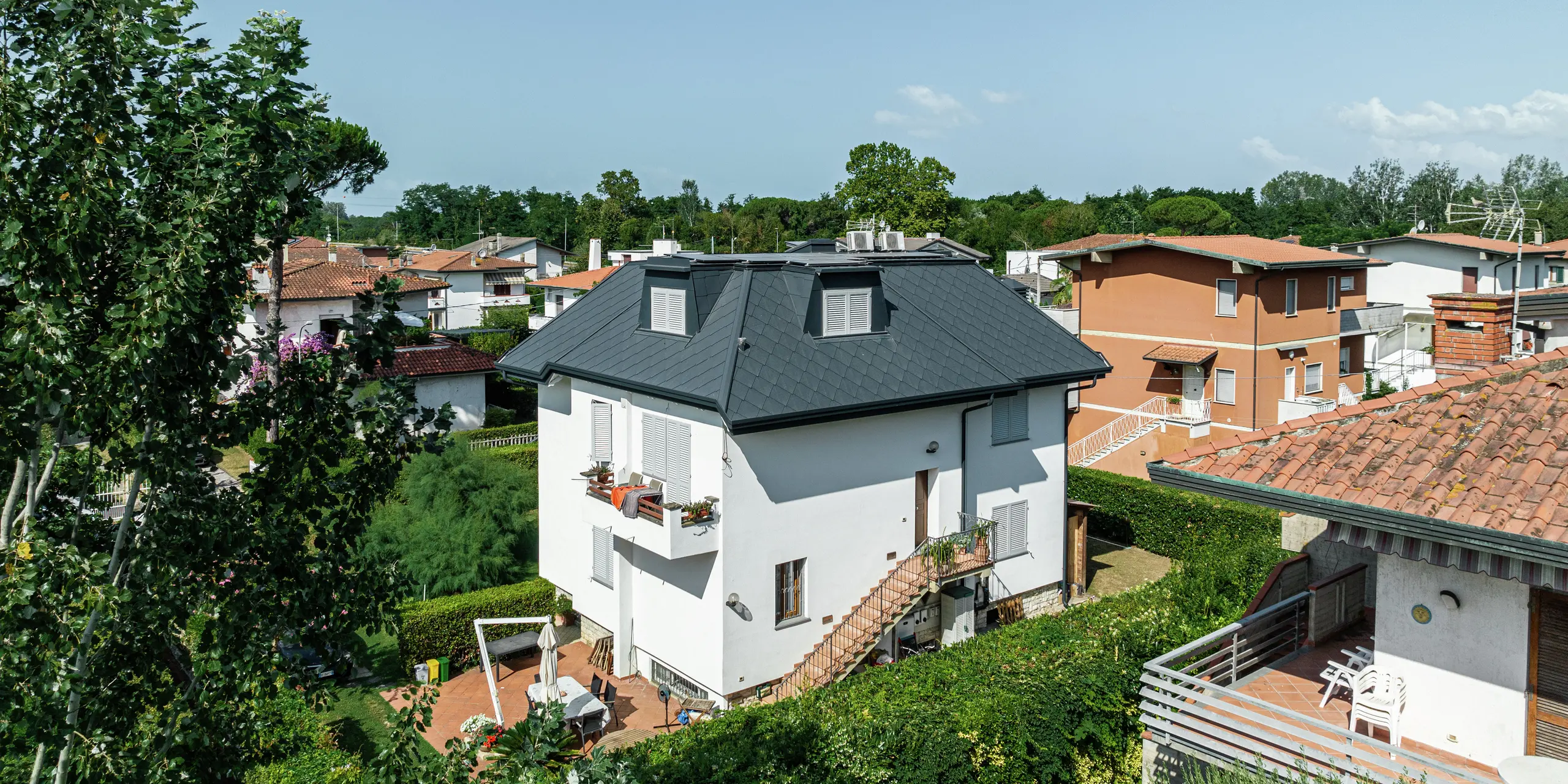 Štýlový rodinný dom v Montignoso z vyvýšenej perspektívy. Strešná šablóna PREFA 44 × 44 vo farbe P.10 kamenná šedá dodáva streche elegantný vzhľad a zaujme svojou trvanlivosťou a extrémnou odolnosťou voči poveternostným vplyvom. Strešný plášť tmavej farby tvorí krásny kontrast k bielej fasáde. Vikiere sú harmonicky integrované do strešnej konštrukcie.