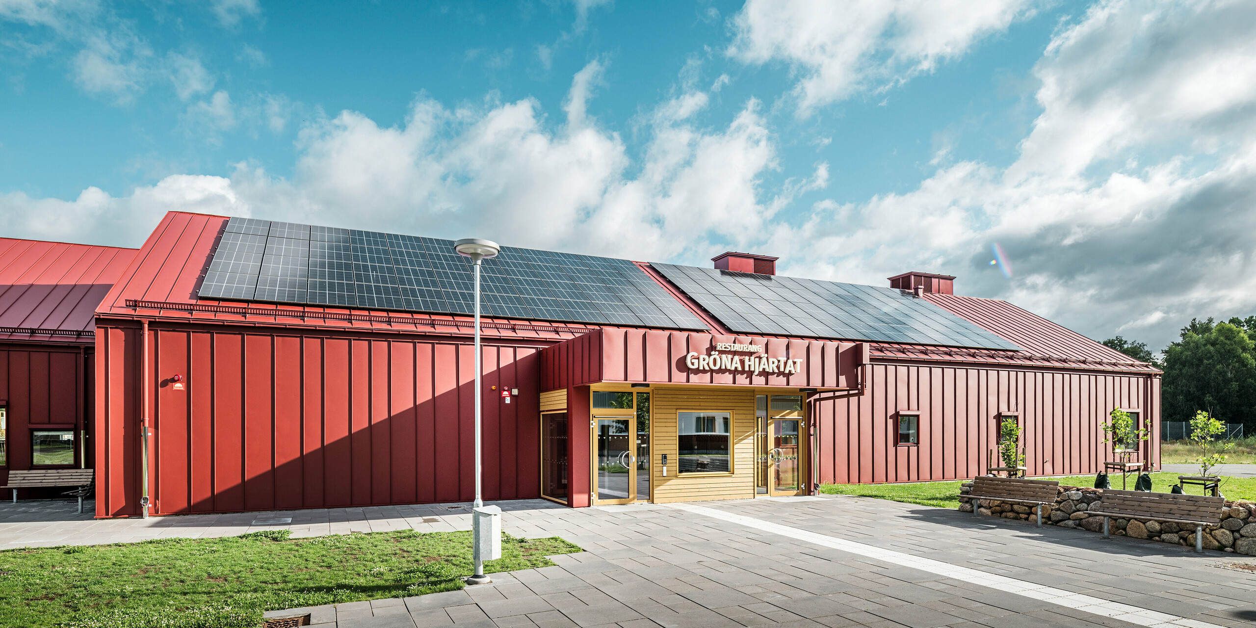 Pohľad na vstupnú časť reštaurácie Gröna Hjärtat, školskej jedálne na Göingeskolan a Prästavångskolan v Broby, Švédsko. Budova dostala pútavú fasádu z materiálu PREFALZ v tmavočervenej farbe. Na streche bol nainštalovaný veľkoplošný fotovoltický systém na zabezpečenie obnoviteľnej energie. Kombinácia odolného hliníkového obkladu a sofistikovaného dizajnu robí z tejto budovy výnimočný príklad modernej architektúry. Strešný odvodňovací systém PREFA má tiež povrchovú úpravu v tmavočervenej farbe, ktorá ladí so strechou a fasádou a podčiarkuje harmonický vzhľad budovy BAuwerk