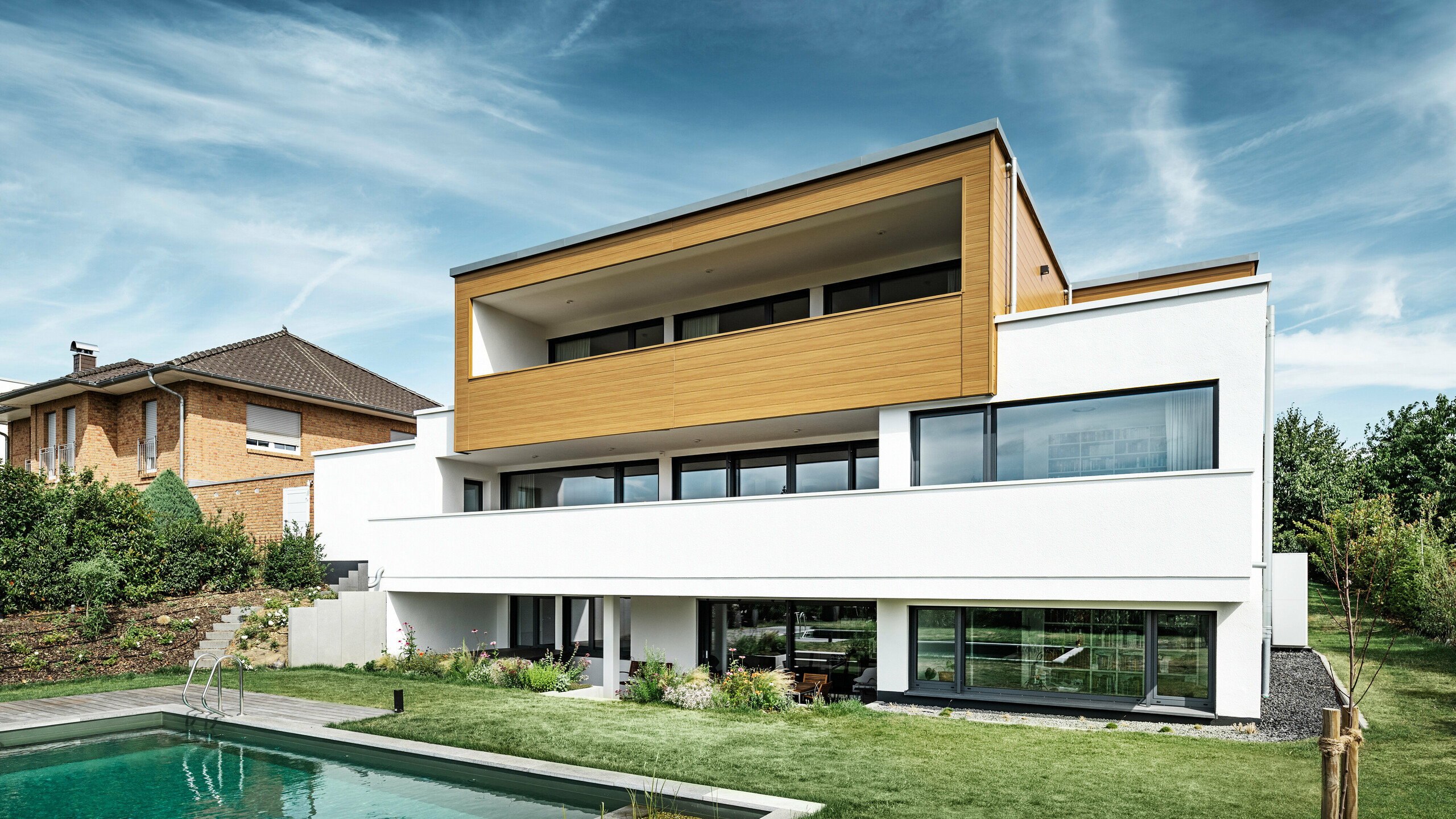 Zadná časť moderného rodinného domu v Usingene, Nemecko. V dolnej časti obrázka je vidieť zelený trávnik, bazén a mladý záhradný strom. Pútavý dom má biely základ budovy z omietnutej fasády, hliníkovú fasádu na hornom poschodí z PREFA Sidings v dub prírodný farbe a veľké okenné plochy s rámom v antracitovej farbe. Z diaľky je na ľavom okraji obrázka vidieť susedný dom s tehlovou fasádou.