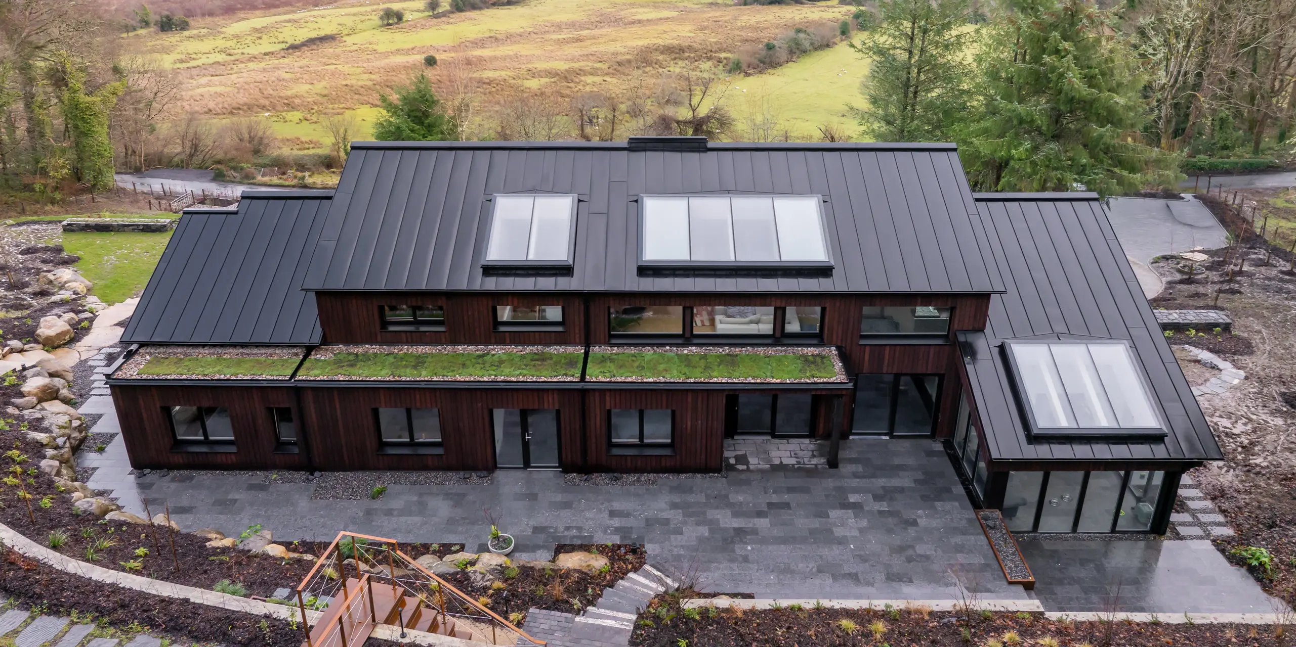 Modernes Einfamilienhaus in Sligo, Irland, mit markanter Holzfassade und elegantem PREFALZ Aluminiumdach in P.10 Schwarz. Die klare Stehfalzstruktur betont die Architektur und fügt sich harmonisch in die weitläufige Landschaft mit Fluss und Hügeln ein. Großzügige Glasflächen und Dachfenster setzen spannende Akzente. Das langlebige, korrosionsbeständige PREFA Dach überzeugt durch Widerstandsfähigkeit gegenüber rauem Klima und sorgt für eine stilvolle, wartungsarme Lösung.