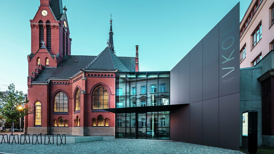 Obrázok zachytáva impozantnú súhru medzi historickým Červeným kostolom a modernou novostavbou v Olomouci. Kostol s charakteristickými gotickými oknami a výraznou vežou tvorí historický chrbtový pilier komplexu, zatiaľ čo moderná prístavba s elegantnou čiernou fasádou z hliníkových kompozitných dosiek PREFABOND predstavuje modernú architektonickú odpoveď. Veľké sklenené plochy umožňujú pohľad do vnútra novej budovy, kde okolie odráža zaujímavú interakciu medzi starým a novým prostredníctvom odrazov v oknách. Presné spracovanie dosiek PREFABOND zdôrazňuje modernú estetiku, zatiaľ čo harmonická integrácia do historického prostredia pôsobí funkčne aj vizuálne.