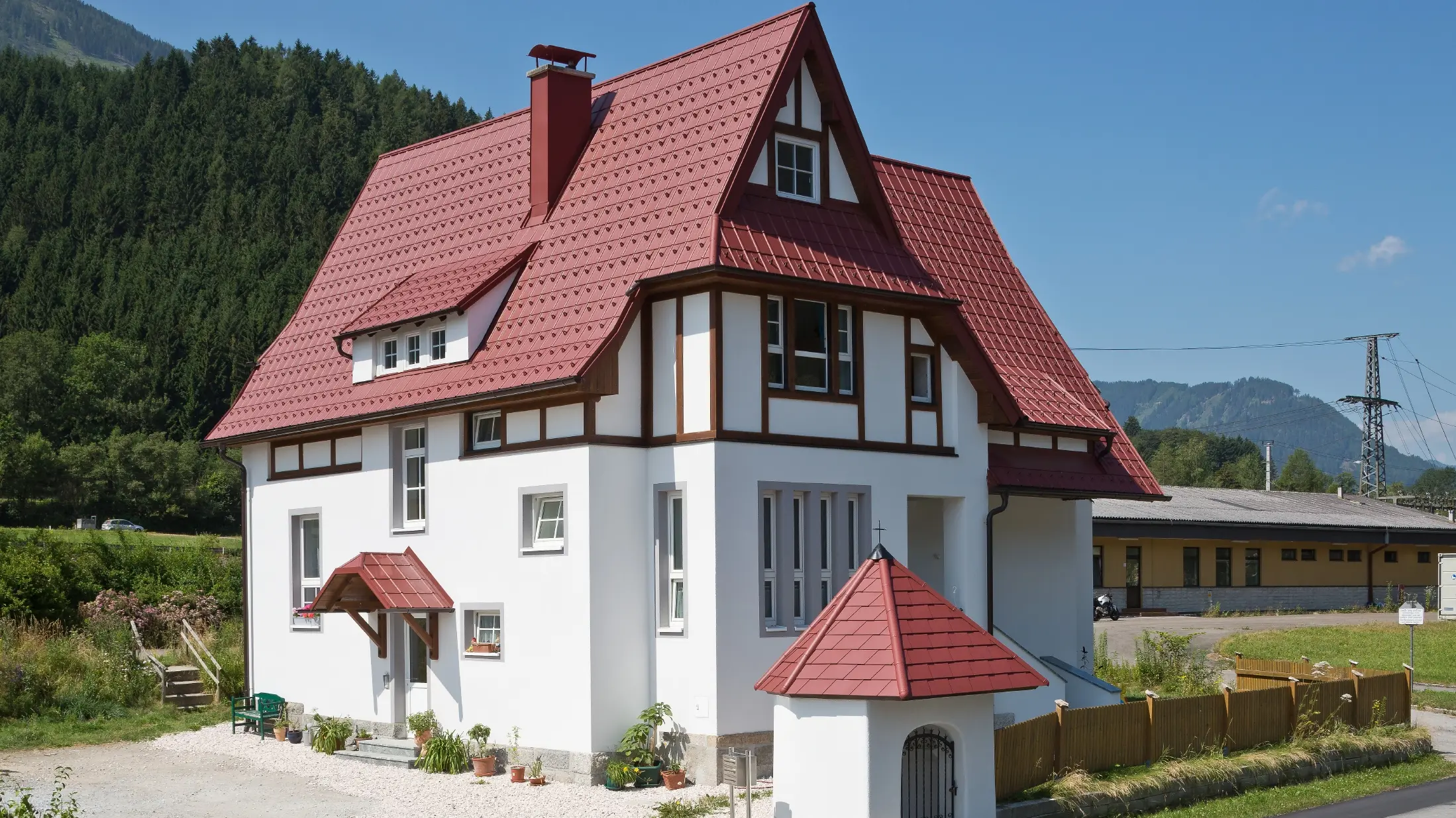 Šikmá strecha so Falcovanou škridlou PREFA v červenej farbe P.10 tmavočervená s mnohými detailmi, ako sú vikiere, vežičky a obruby komínov. Prístrešok a kaplnka pred domom sú tiež pokryté plechovou krytinou PREFA.